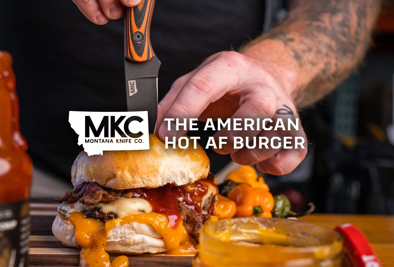 A close-up view of a man’s tattooed hands holding a burger with a knife stuck into it.