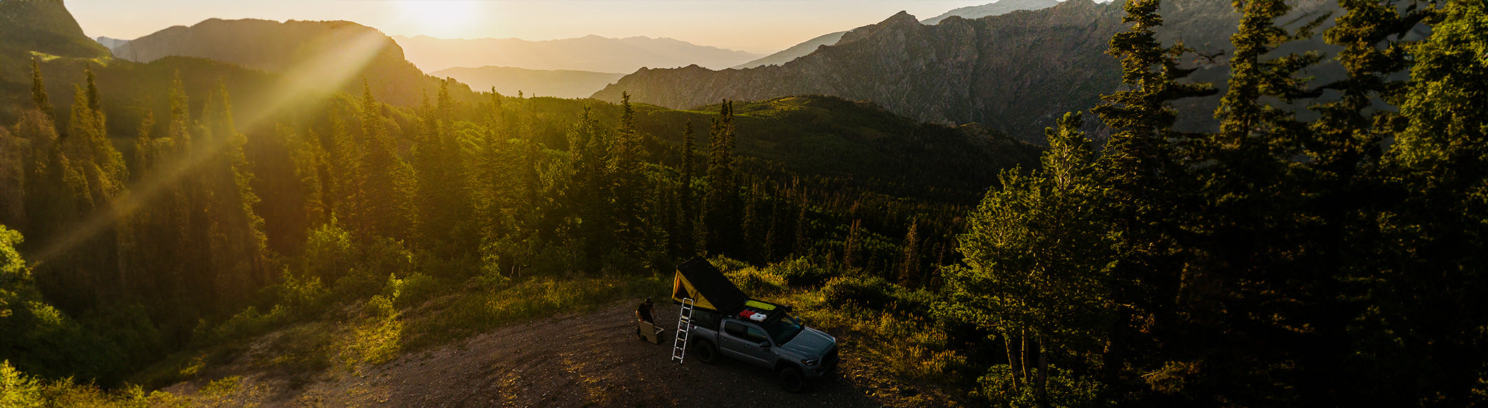 Scenic mountain view at sunset with a vehicle and camping setup among tall trees, perfect for hiking and camping adventures.