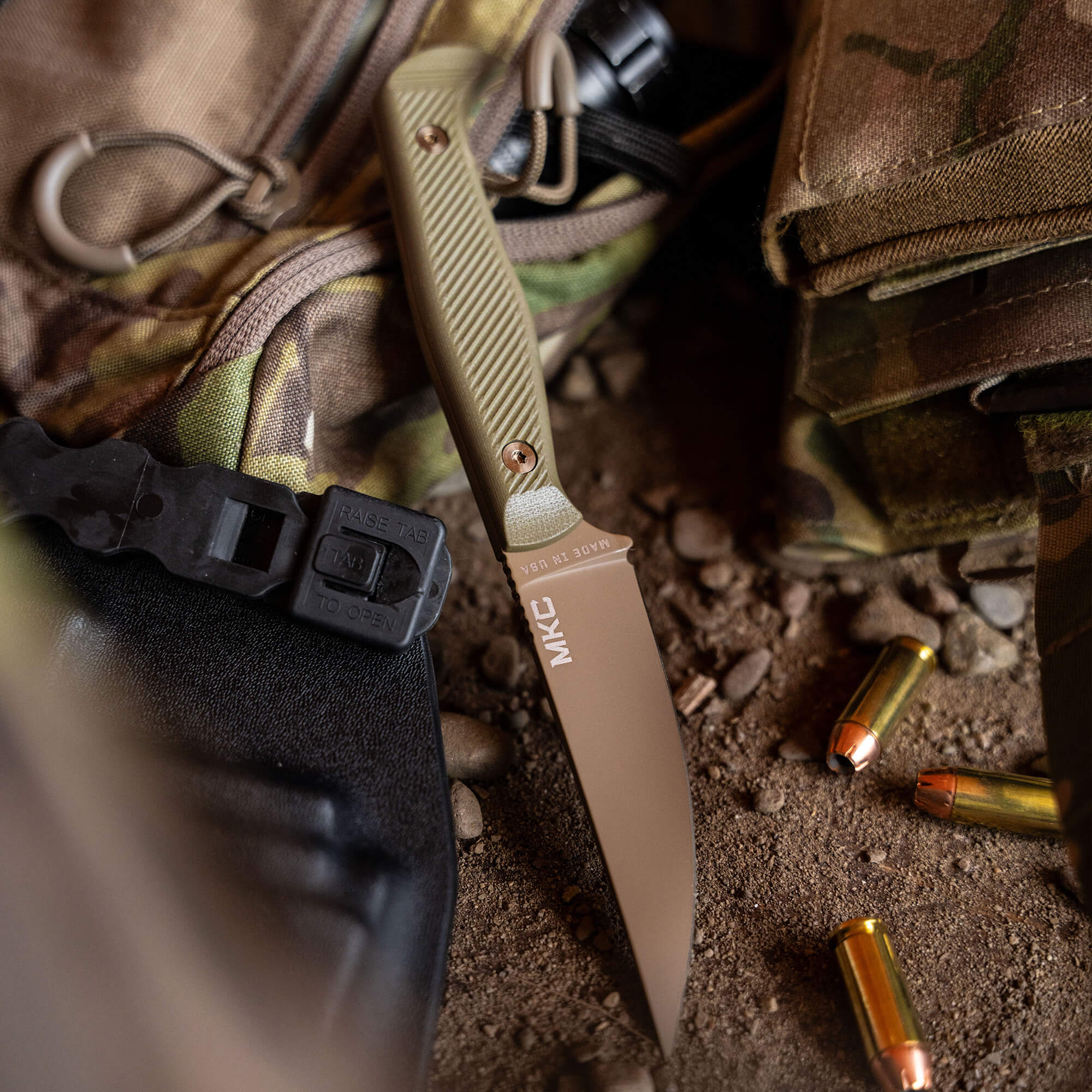 Alt text: "Close-up of a BATTLE GOAT coyote olive drab tactical knife with MKC logo, positioned against a tactical backpack and military equipment, with scattered bullet casings on a gritty surface."