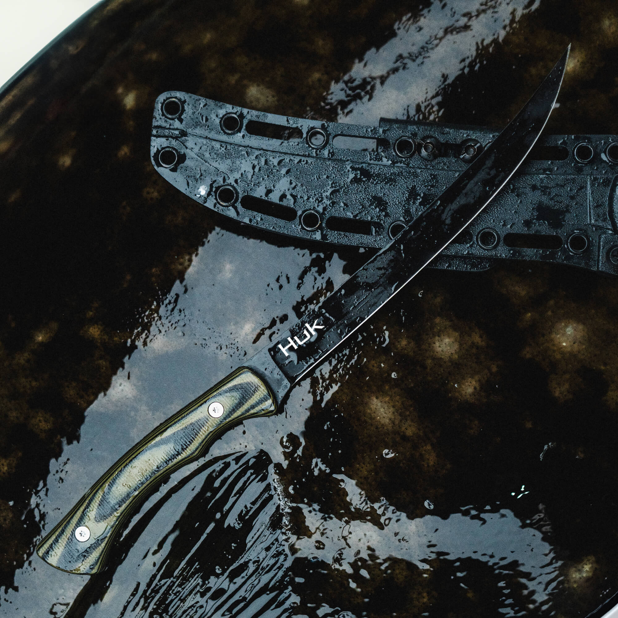 HUK Flathead Fillet knife with a sleek black blade and textured handle, resting on a wet surface.