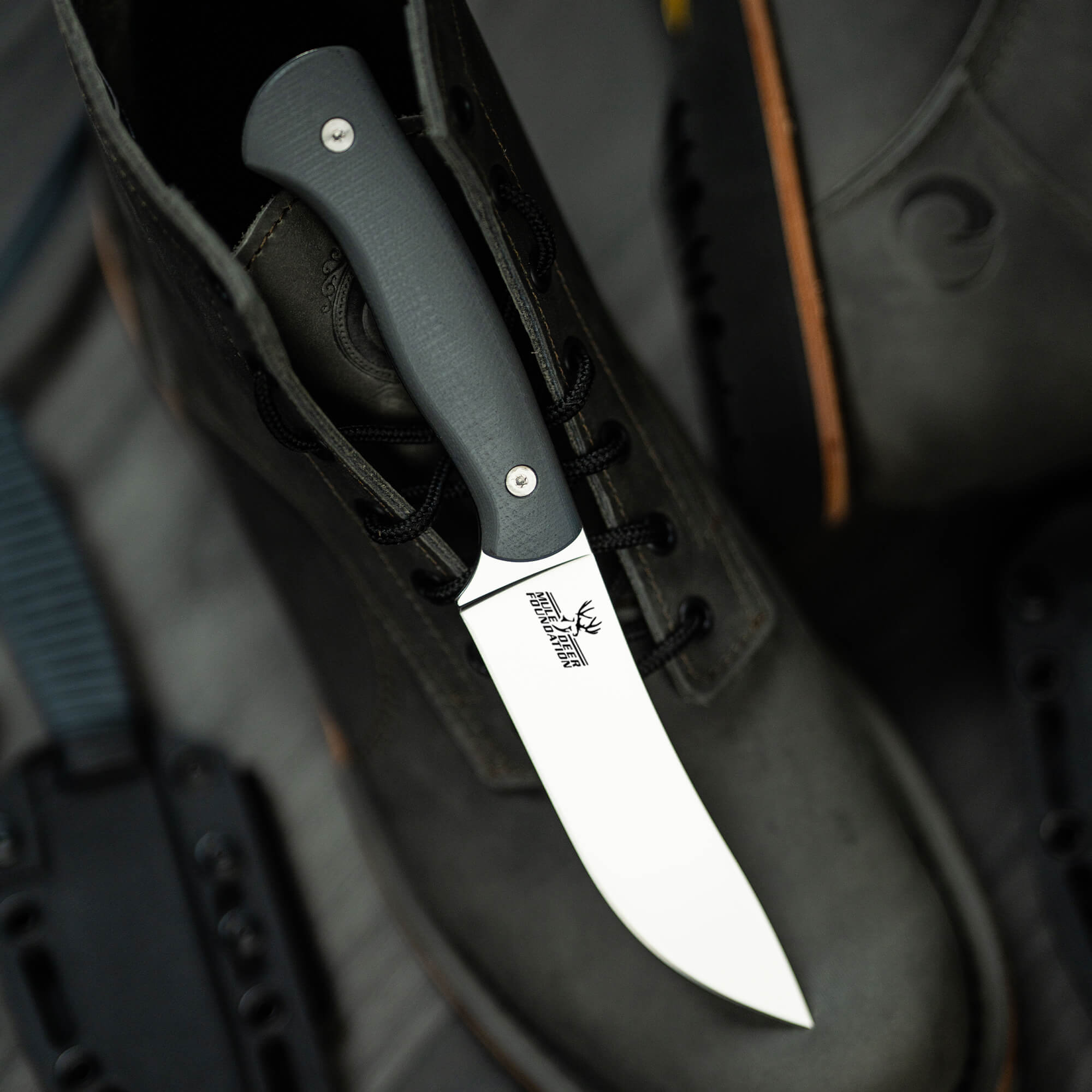Close-up image of a MagnaCut Stonewall Skinner knife by Mule Deer Foundation with a grey handle, displayed tucked into a black boot emphasizing its sleek design and sharp blade, featuring the foundation's logo on the blade.