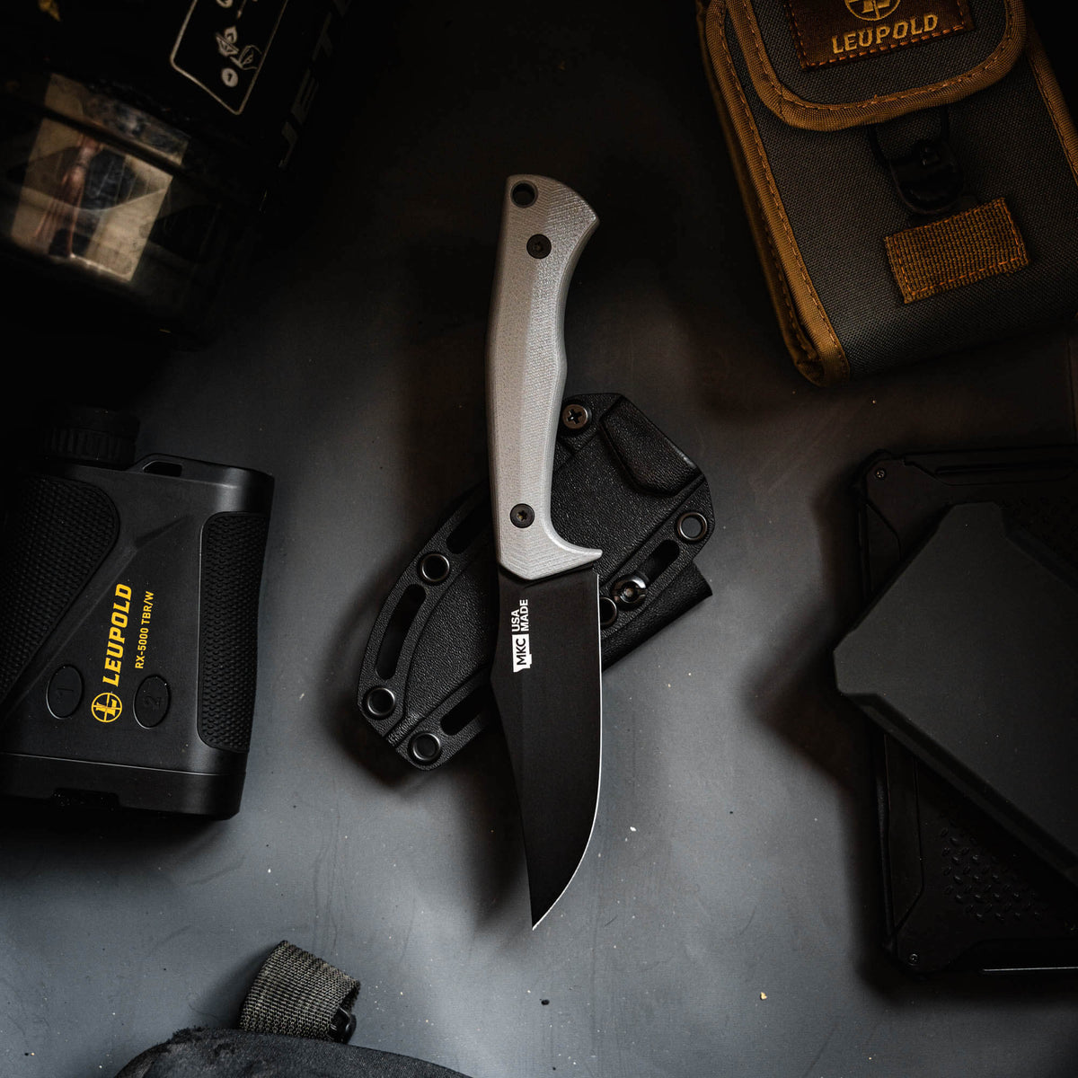 Triumph Pro grey fixed-blade knife on a dark surface surrounded by tactical gear and accessories.