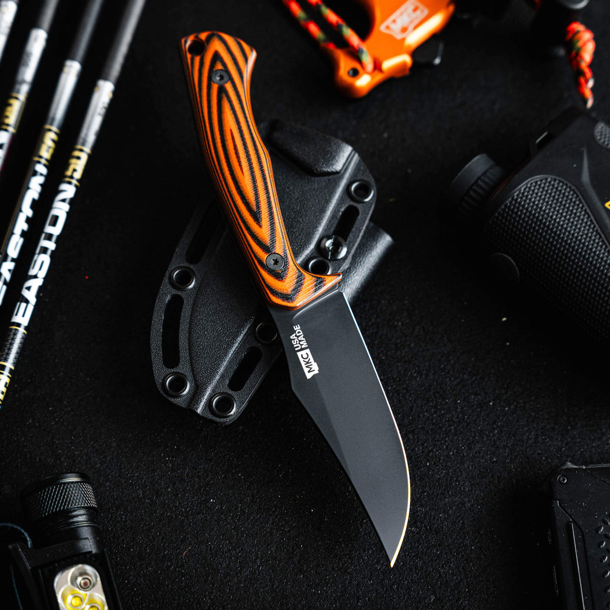 Triumph Pro knife in orange and black with a sleek blade, displayed on a black background among outdoor gear.