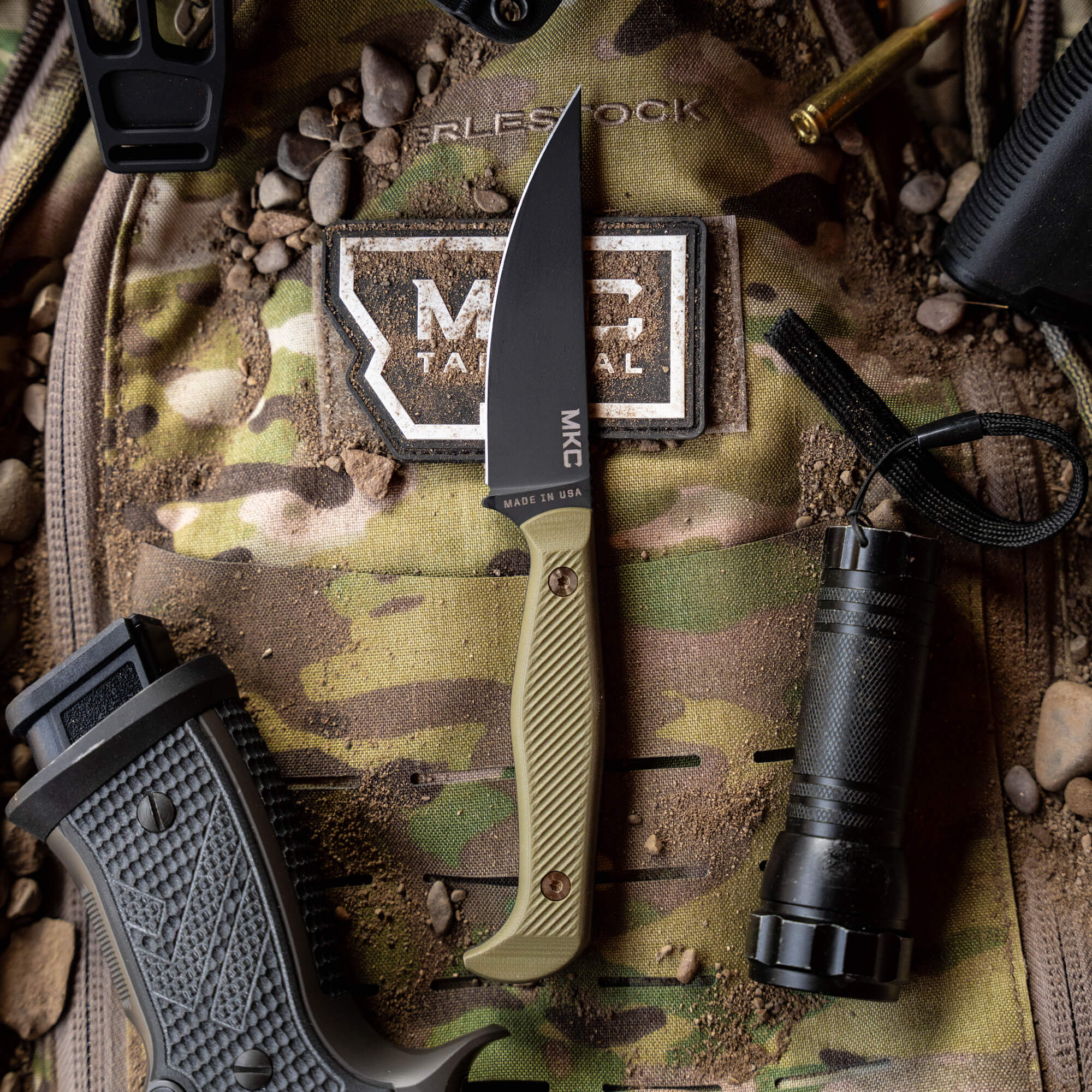 Alt text: "Close-up view of the Battle Goat tactical knife with a black blade and olive drab handle, displayed on a camouflage background surrounded by tactical gear including a flashlight and a holster."