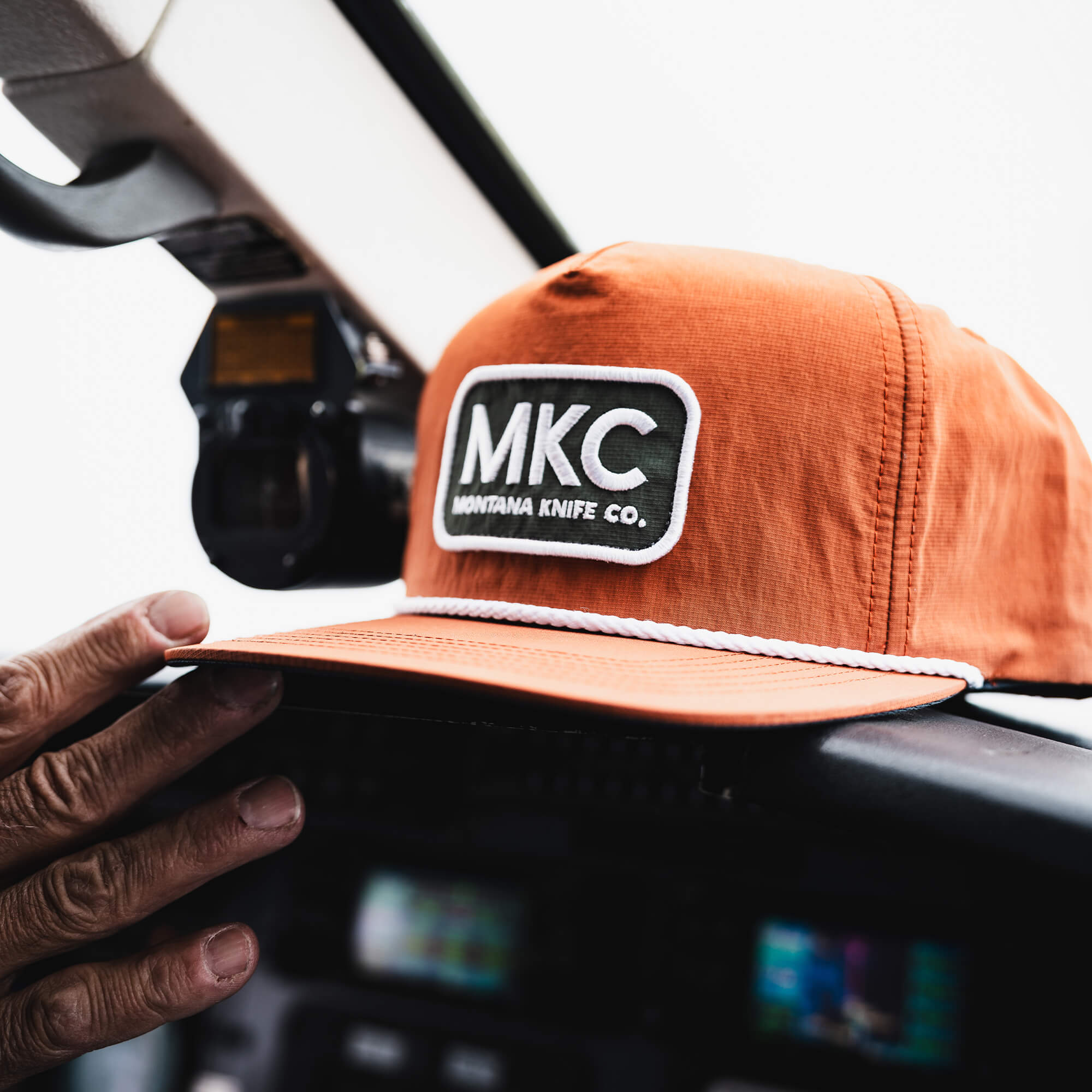 Rust-colored MKC River Rope Hat with logo, resting on a cockpit dashboard, showcasing outdoor style and functionality.