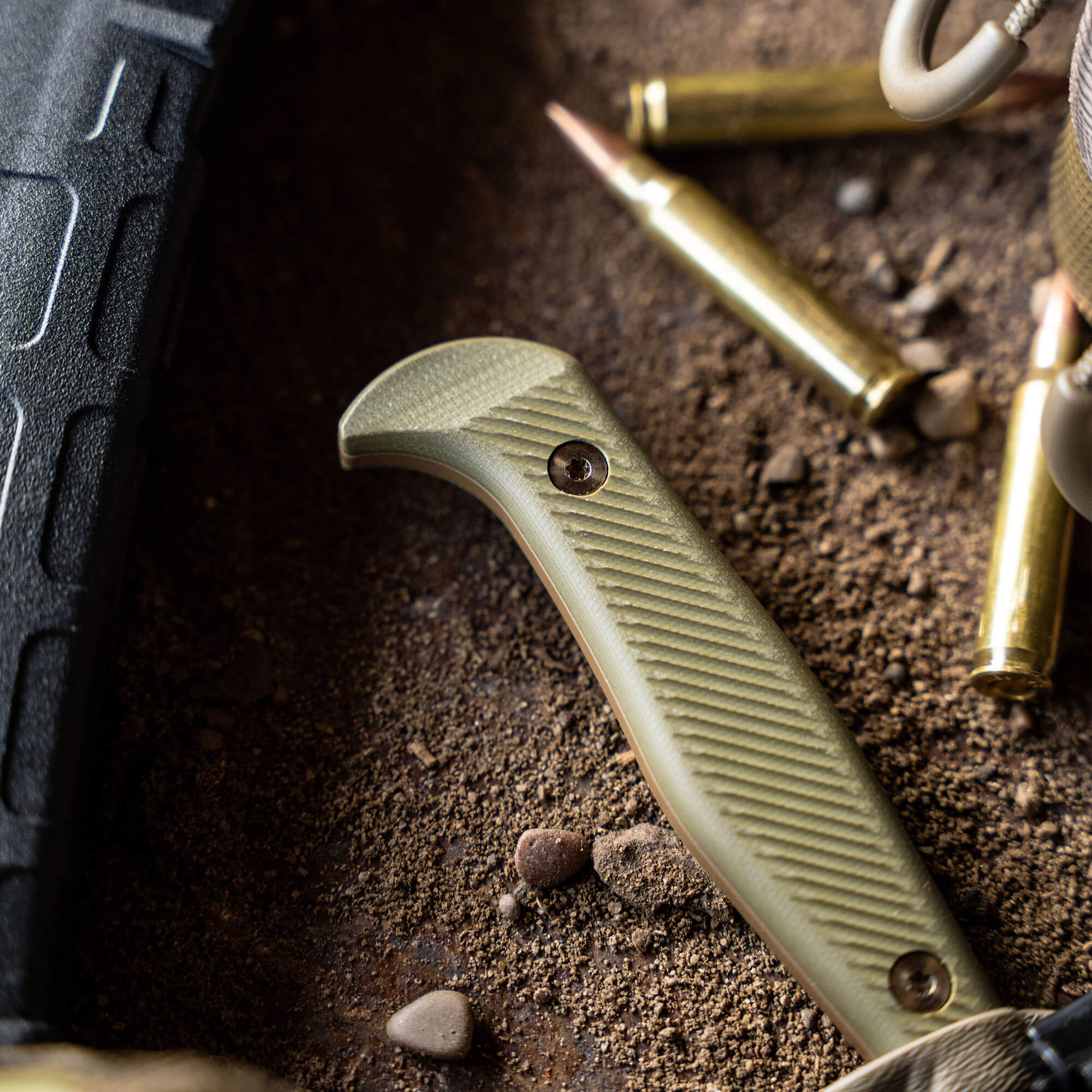 Close-up view of a coyote olive drab BATTLE GOAT tactical knife handle with textured grip, surrounded by scattered ammunition and earthy terrain.