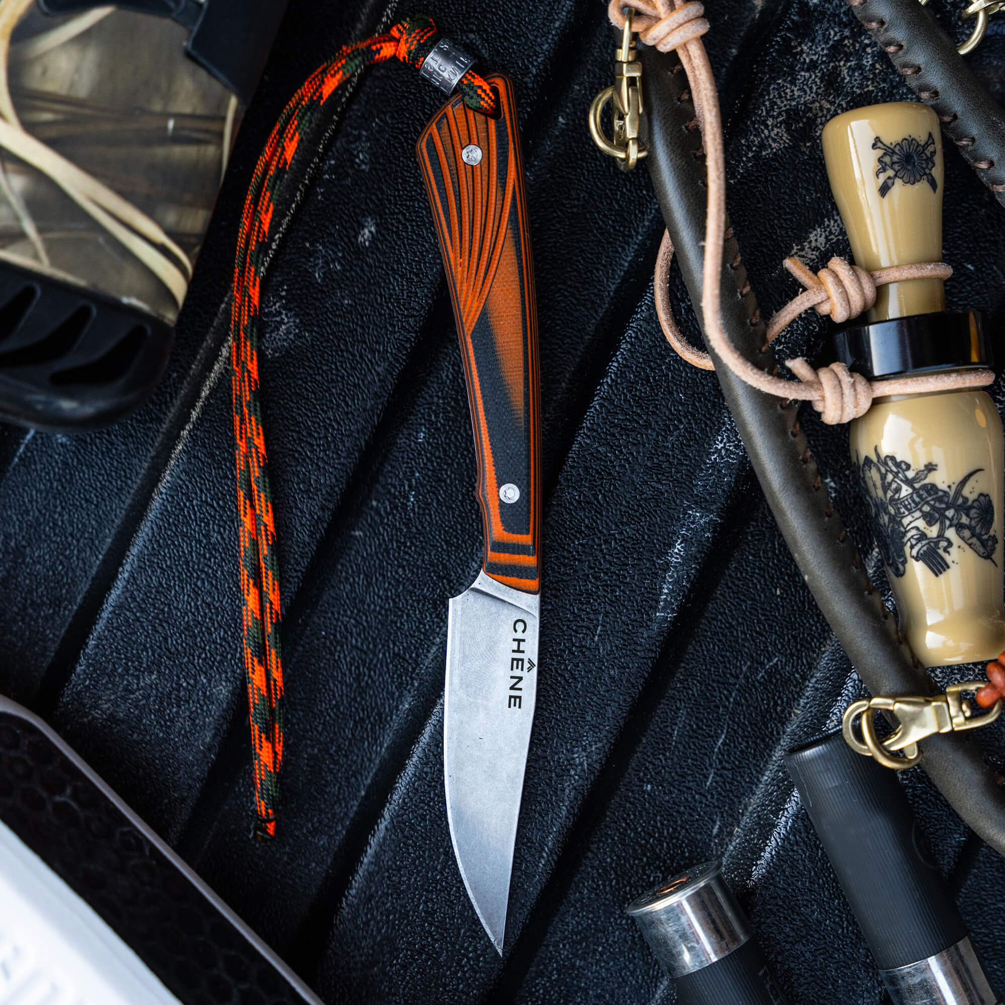 CHÊNE Freezout collaboration knife with a sleek blade and striking orange and black handle, surrounded by outdoor gear.