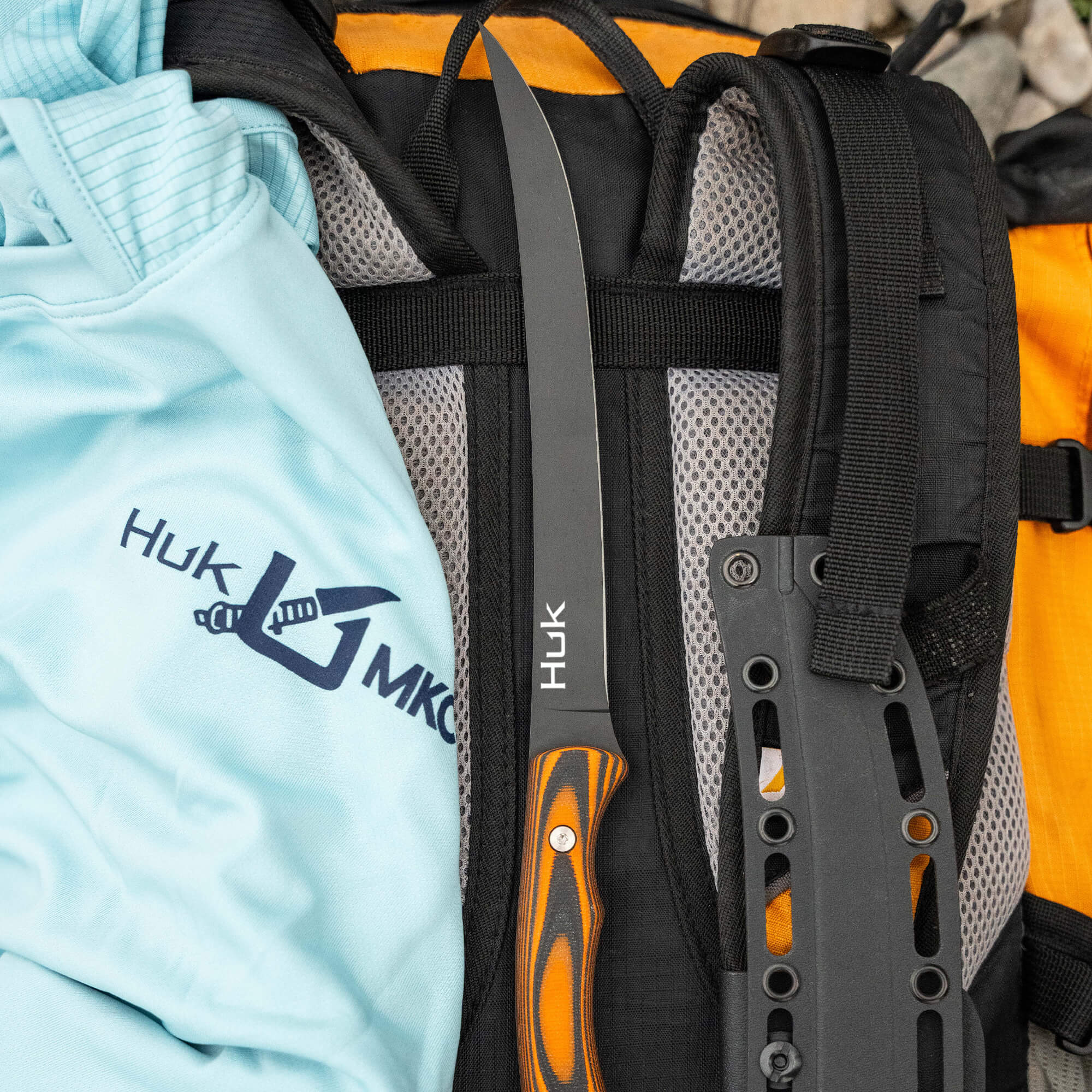 HUK Flathead Fillet knife resting on a backpack beside a light blue shirt, showcasing its sleek design and functionality.