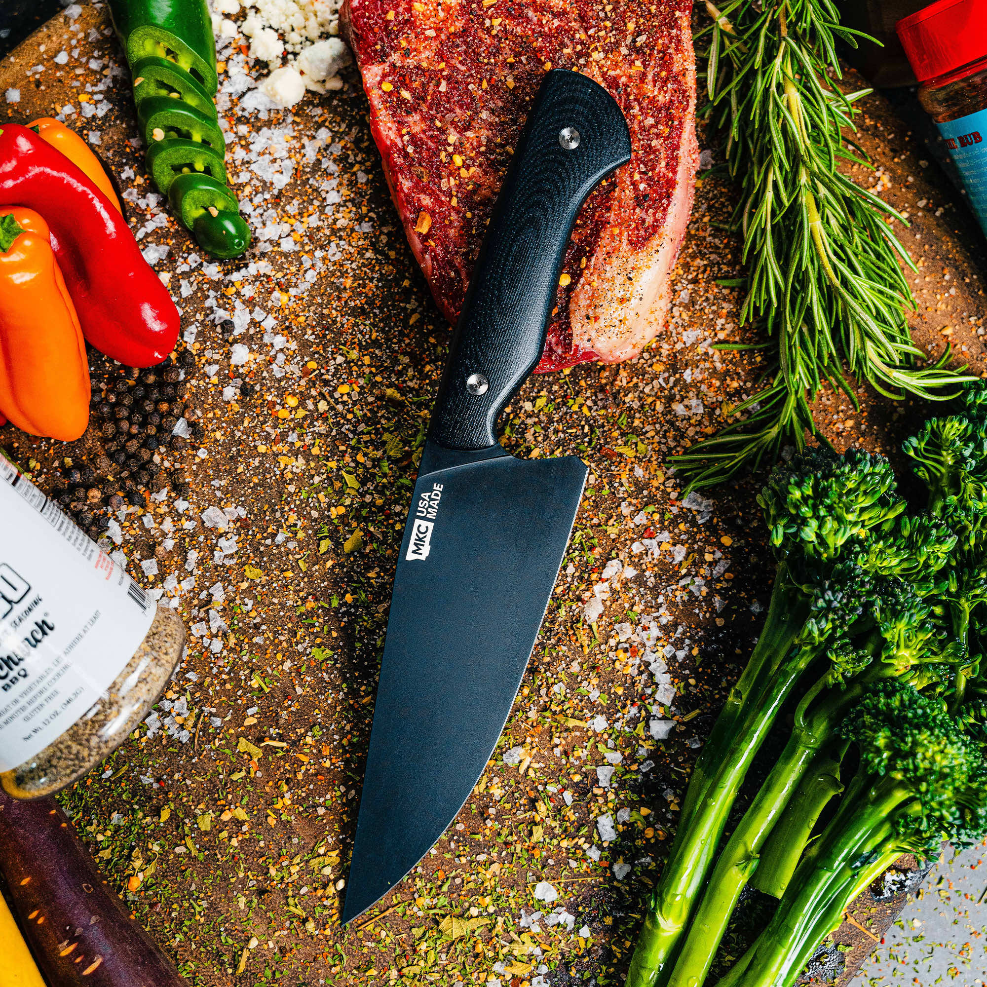 Meat Church chef knife on a rustic cutting board surrounded by fresh vegetables and spices, showcasing its sleek design.