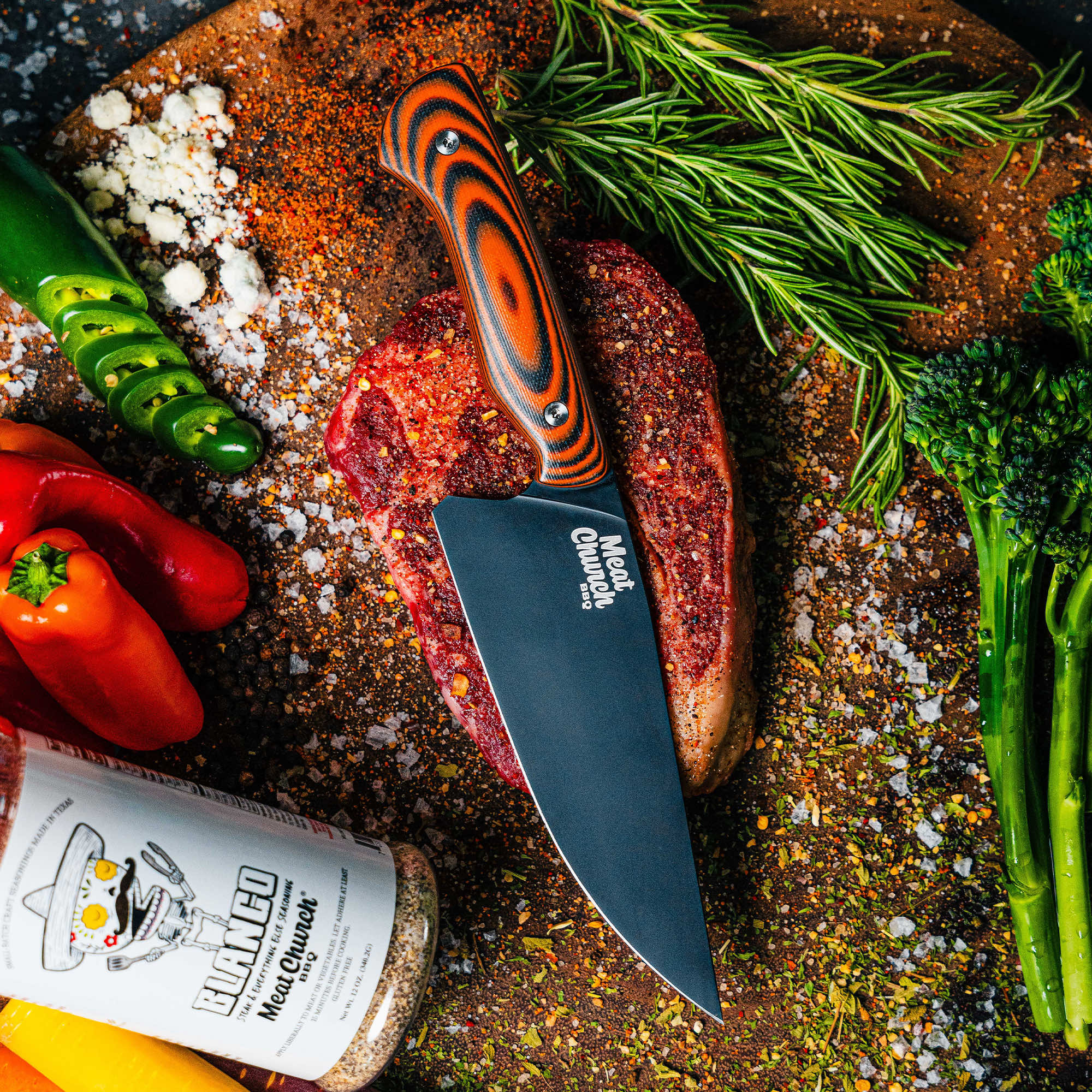 Meat Church chef knife on a cutting board with seasoned steak and fresh vegetables, showcasing its sleek design and functionality.