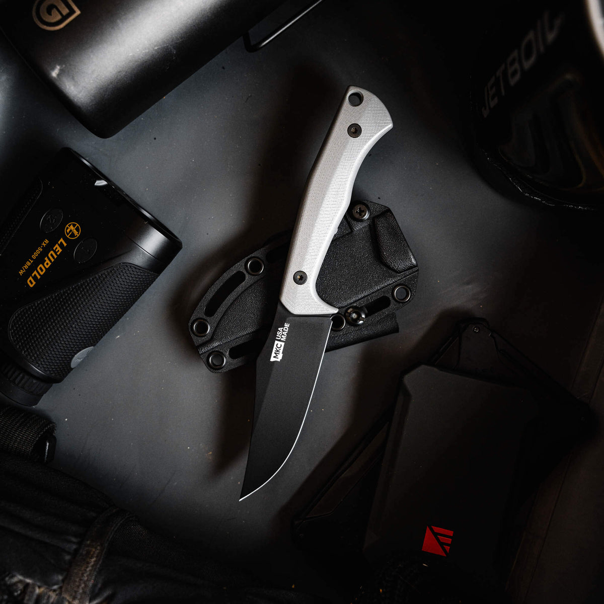 Grey Triumph Pro folding knife with a black blade, placed on a dark surface among outdoor gear.