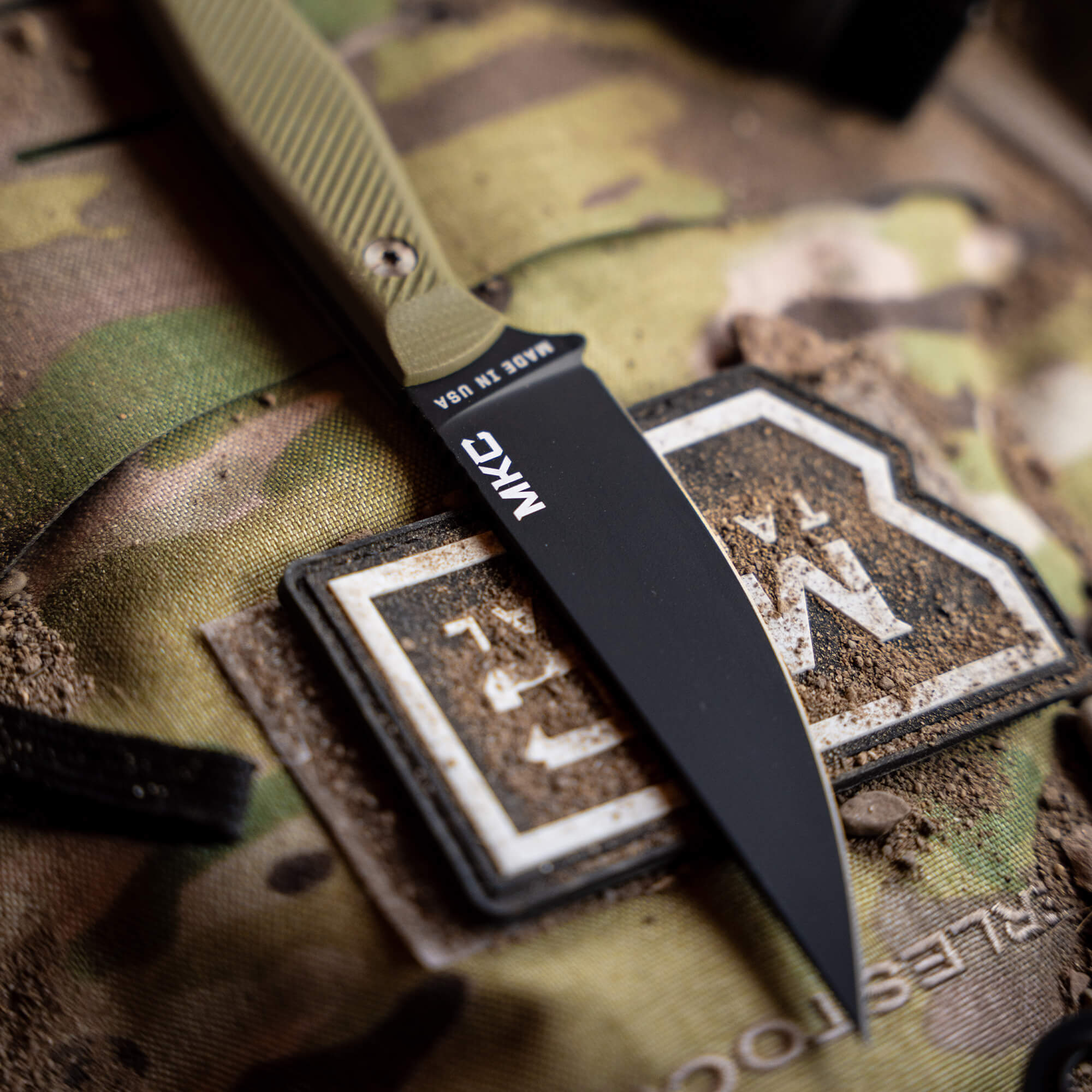 Close-up of a black tactical knife, MKC Battle Goat - BLK/OD, with olive drab handle lying on a camouflage surface, focused on blade's detail and branding.