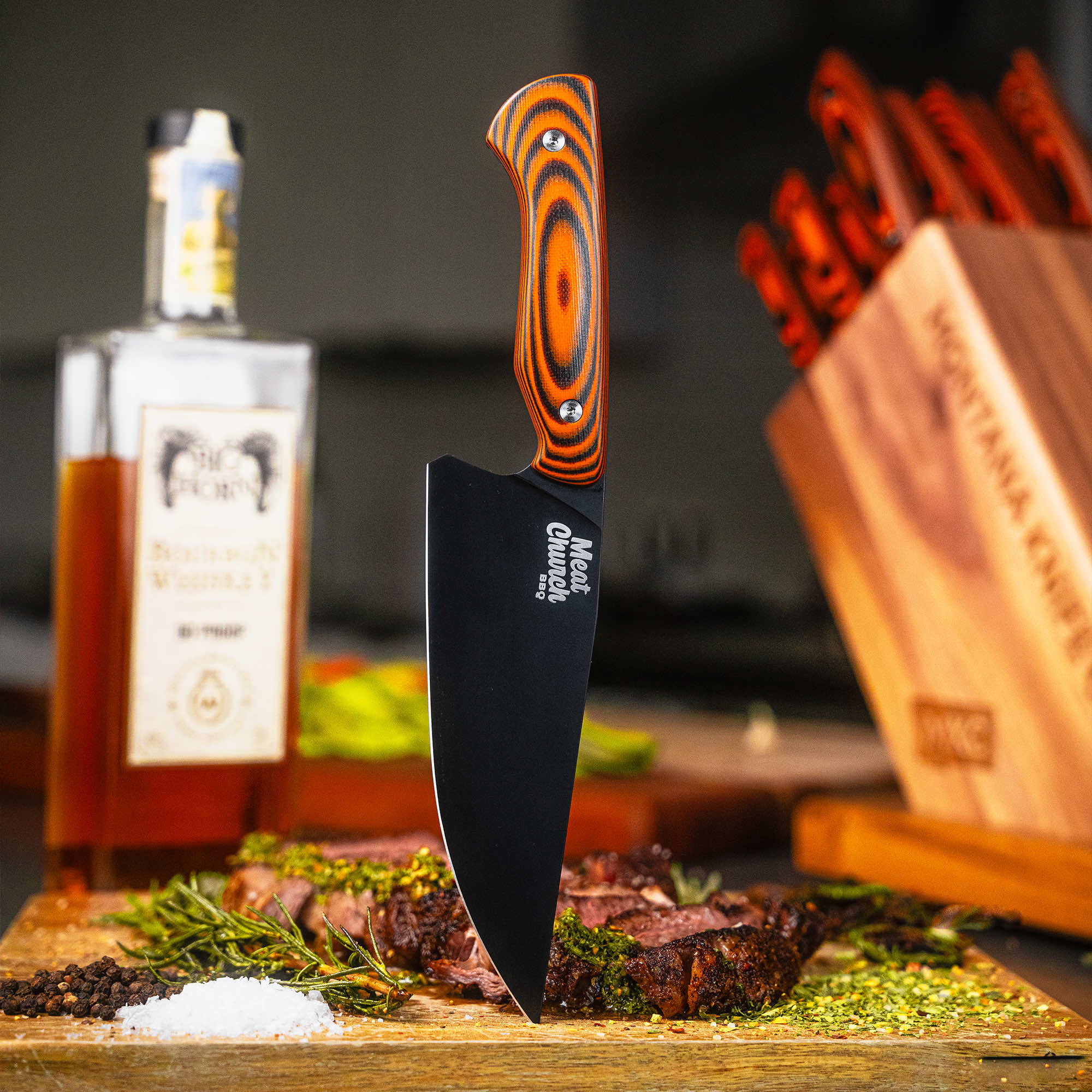 Montana Knife Company's Meat Church Chef Knife on a cutting board with seasoned meat, whiskey bottle in the background.