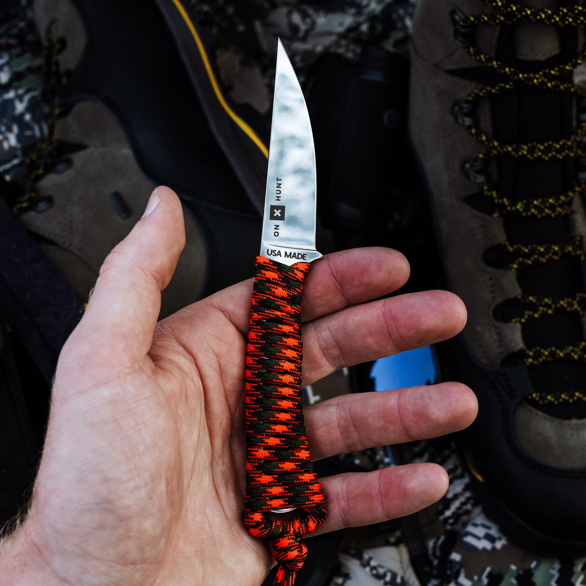 ONX Hunt Mini-Speedgoat 2.0 knife held in hand, featuring a sharp blade and paracord handle, surrounded by outdoor gear.