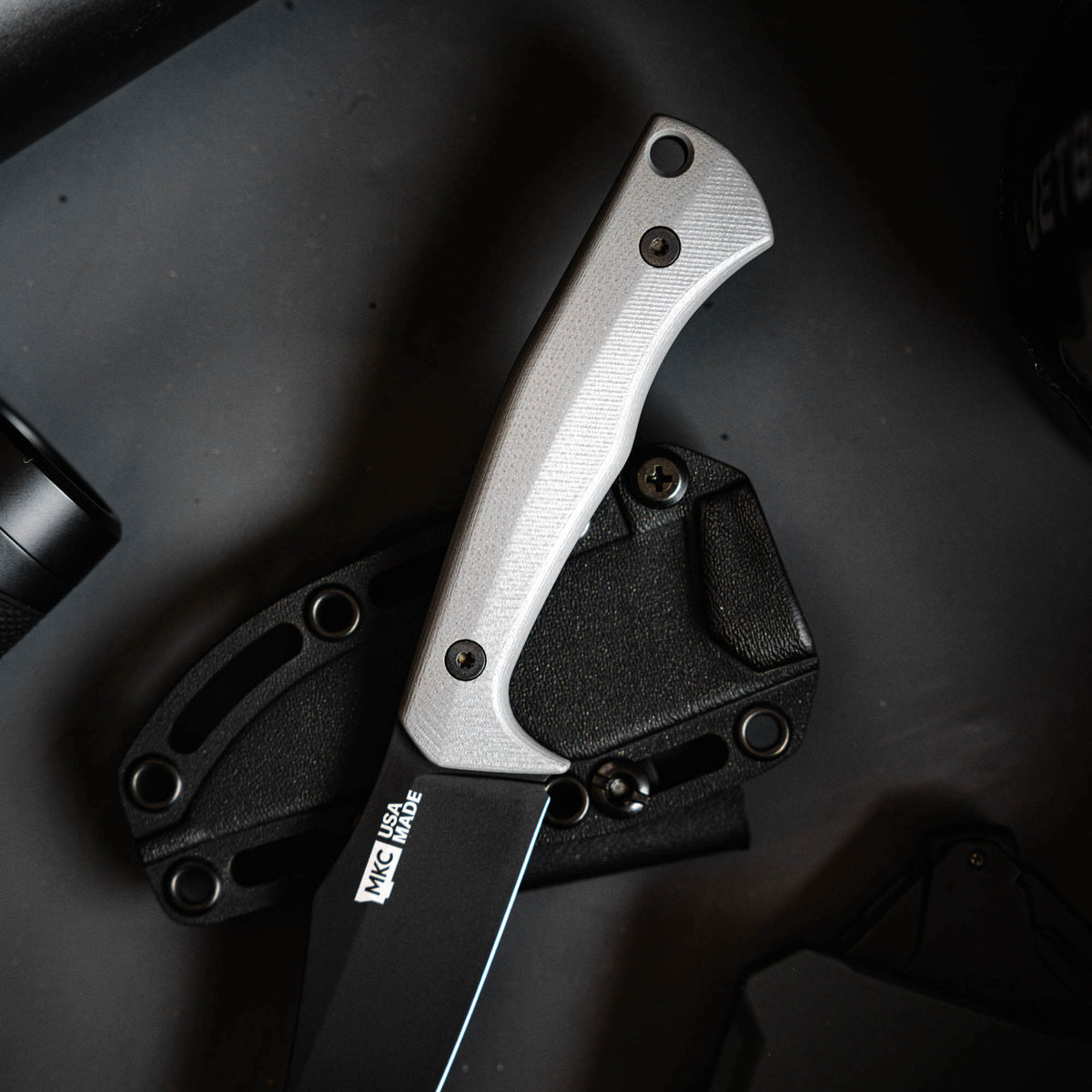 Triumph Pro knife in grey with textured handle, displayed on a dark surface alongside a sheath and tools.