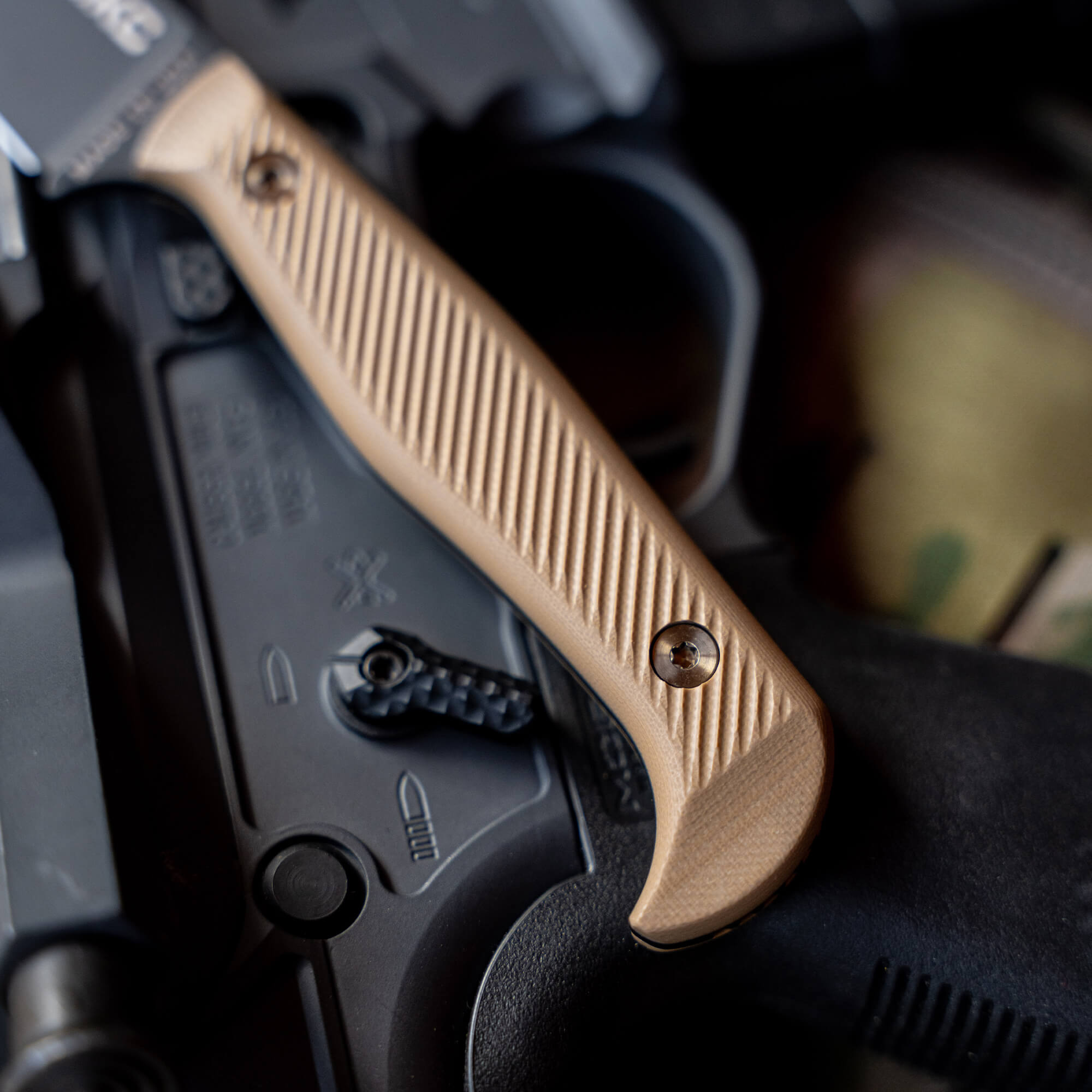 Close-up of a tan-colored tactical knife, the BATTLE GOAT in BLK/COY, resting on a dark, textured surface with firearm equipment partially visible under the handle.