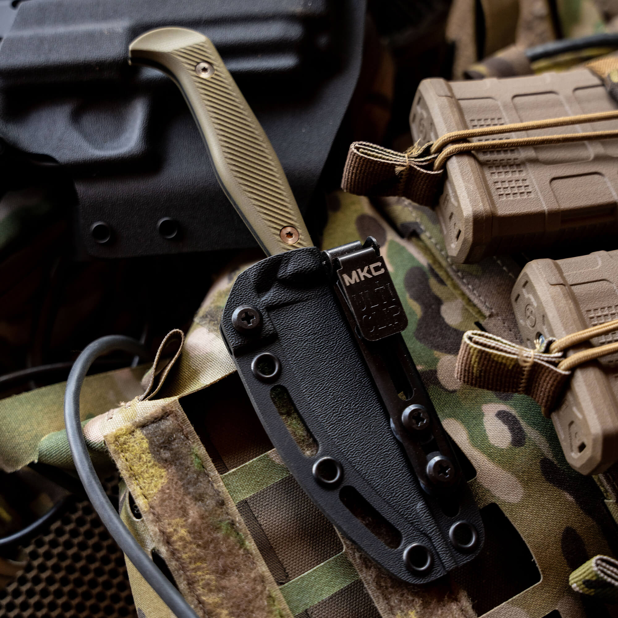 Alt text: Close-up of a black Battle Goat tactical knife in a secure sheath, displayed with additional military gear and camouflage equipment in the background, emphasizing the knife's rugged design and utility in tactical environments.