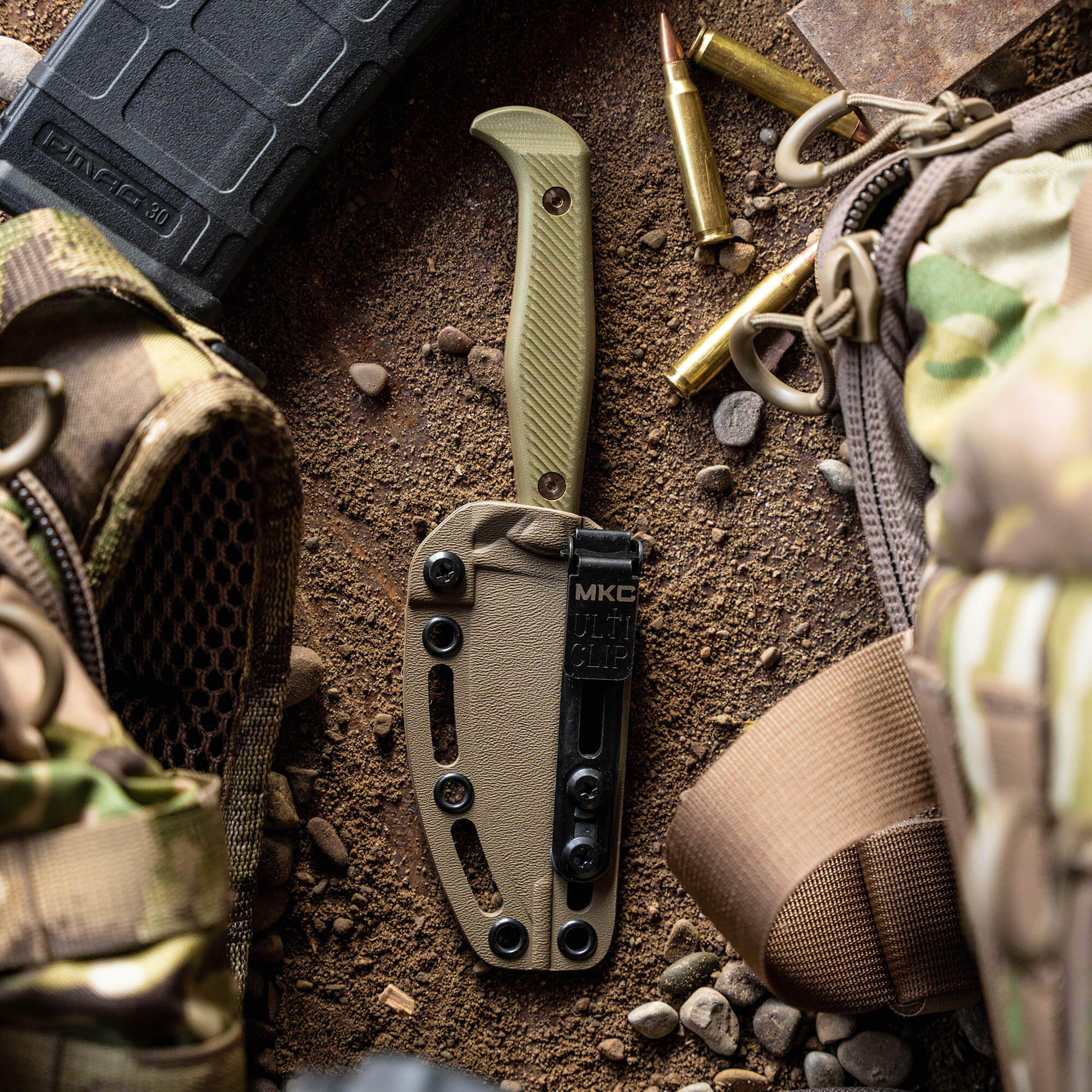 Alt text: "Close-up of a BATTLE GOAT tactical knife in Coyote/Olive Drab with a textured handle and black multi-clip sheath, displayed on a background of tactical gear, spent ammunition, and sandy terrain."