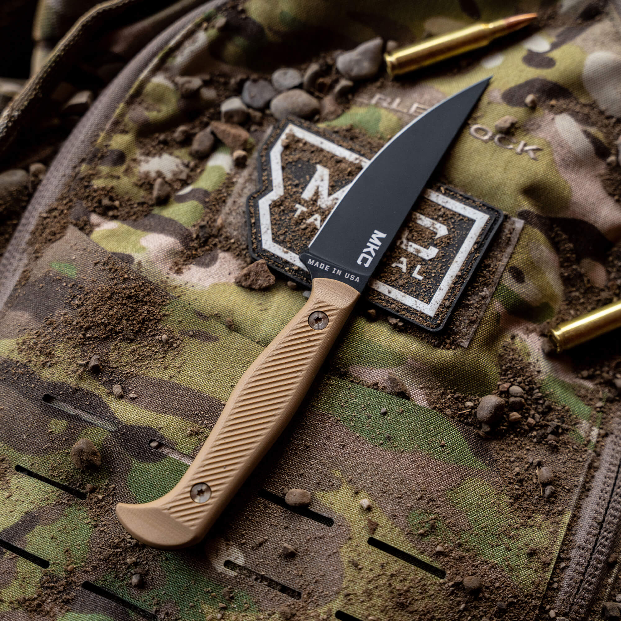 Alt text: "Close-up of a Battle Goat fixed-blade knife with a black blade and coyote tan handle, resting on a camouflaged backpack decorated with patches and surrounded by spent bullet casings, highlighting rugged outdoor use."