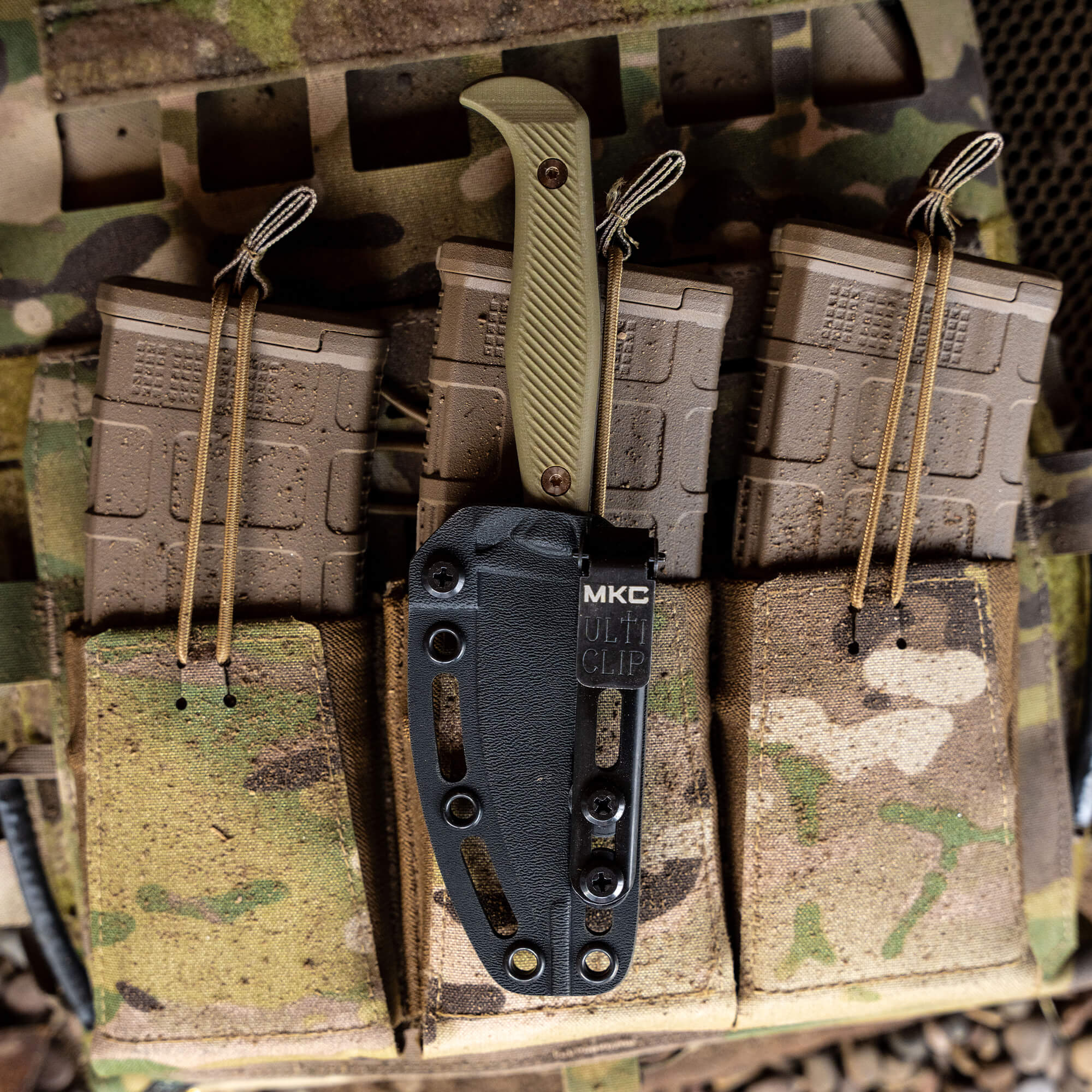Alt text: "Close-up view of the BATTLE GOAT survival knife in olive drab, attached to a camouflage tactical vest with multi-clip mount. The knife is stored in a black sheath and positioned next to several tactical pouches on