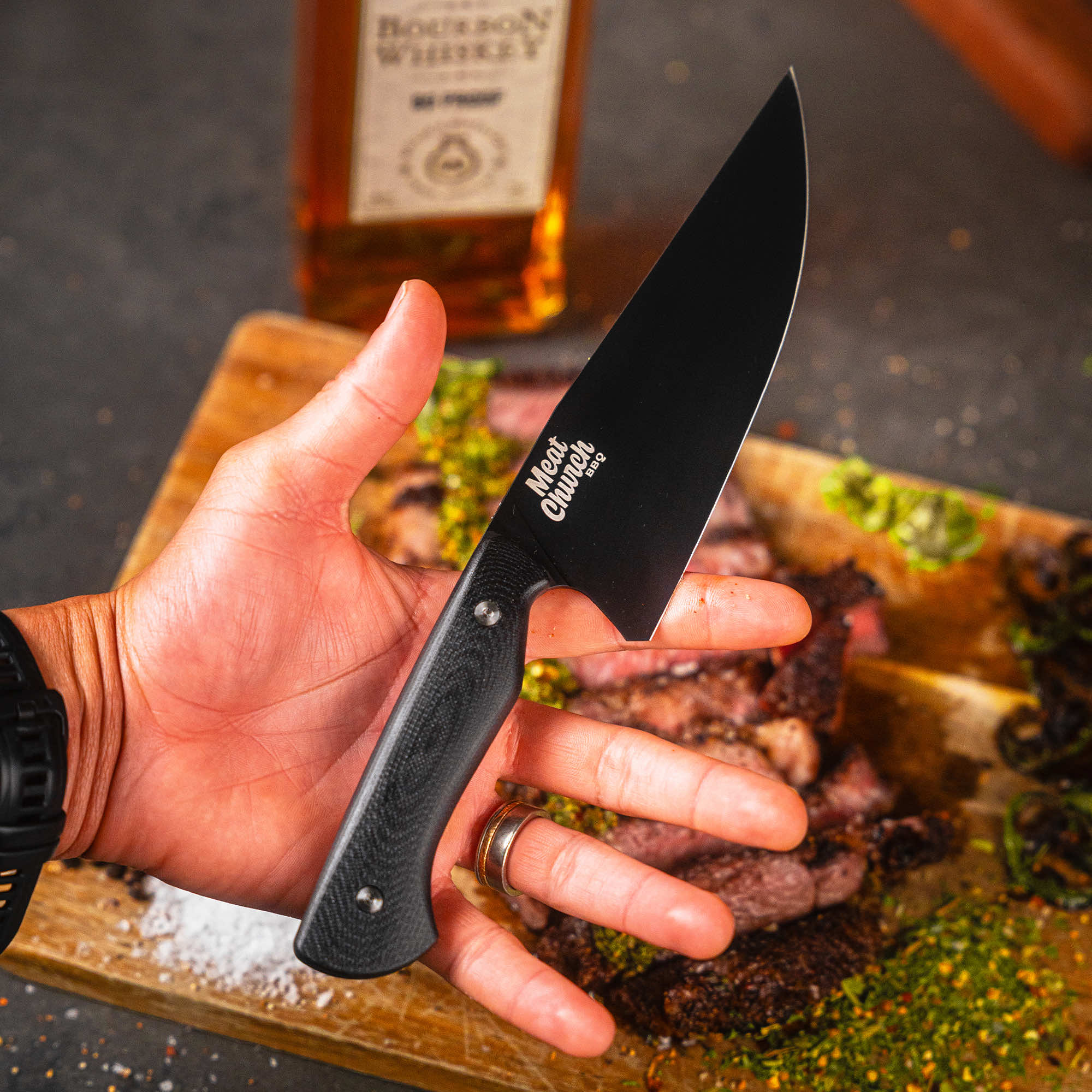 Montana Knife Company Meat Church chef knife held in hand, showcasing its sleek design against a backdrop of grilled meat.