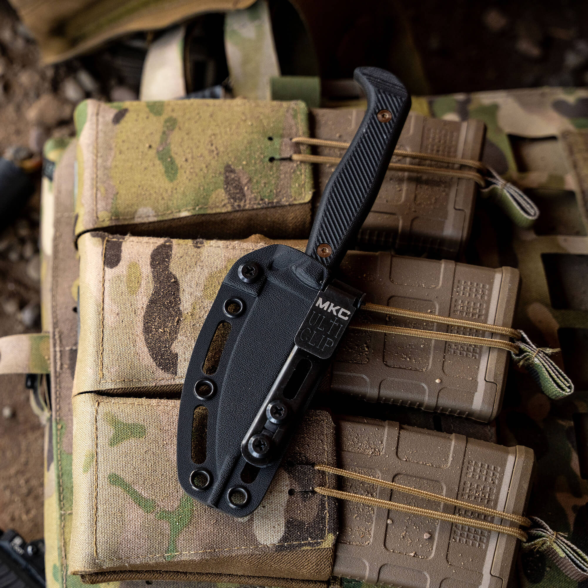 Alt text: "Black Battle Goat tactical knife with a textured handle and black sheath, attached to a multicam tactical vest alongside camouflage ammunition pouches."