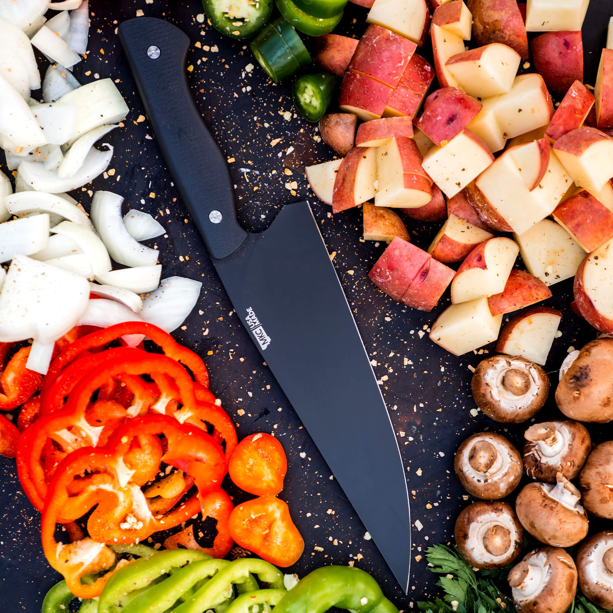 Sharp Bighorn Chef black knife lying on a dark surface surrounded by colorful chopped vegetables including bell peppers, onions, potatoes, and mushrooms, perfectly demonstrating the product's utility in food preparation.