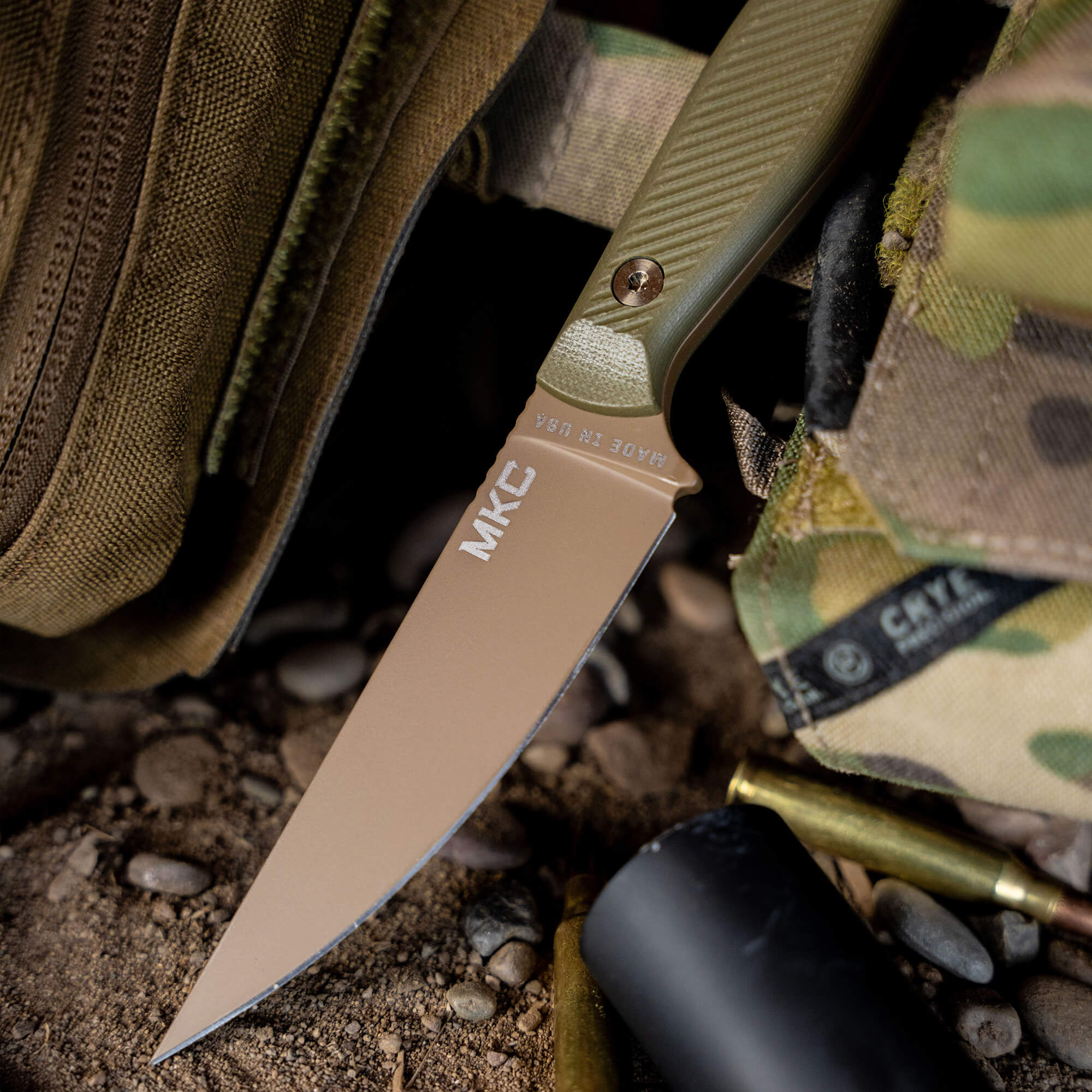 Alt text: "Close-up of a Battle Goat tactical knife in coyote brown and olive drab handle, protruding from a rugged outdoor setting with pebbles, a backpack, and camouflage material, emphasizing its robust and durable design suitable for military