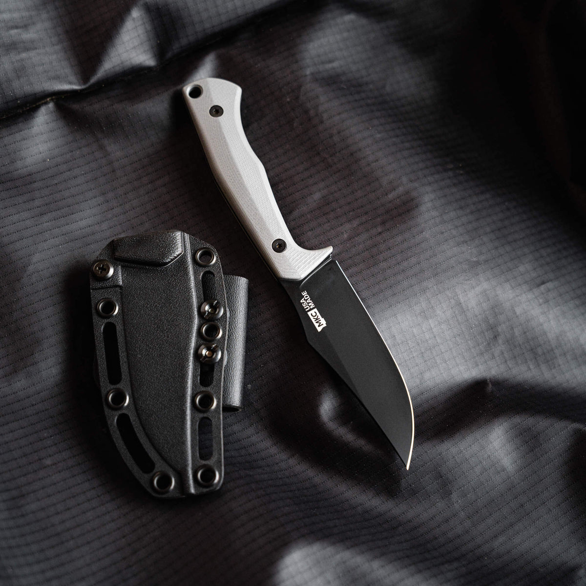 Triumph Pro Grey knife with a black blade and grey handle, displayed alongside its black sheath on a textured surface.