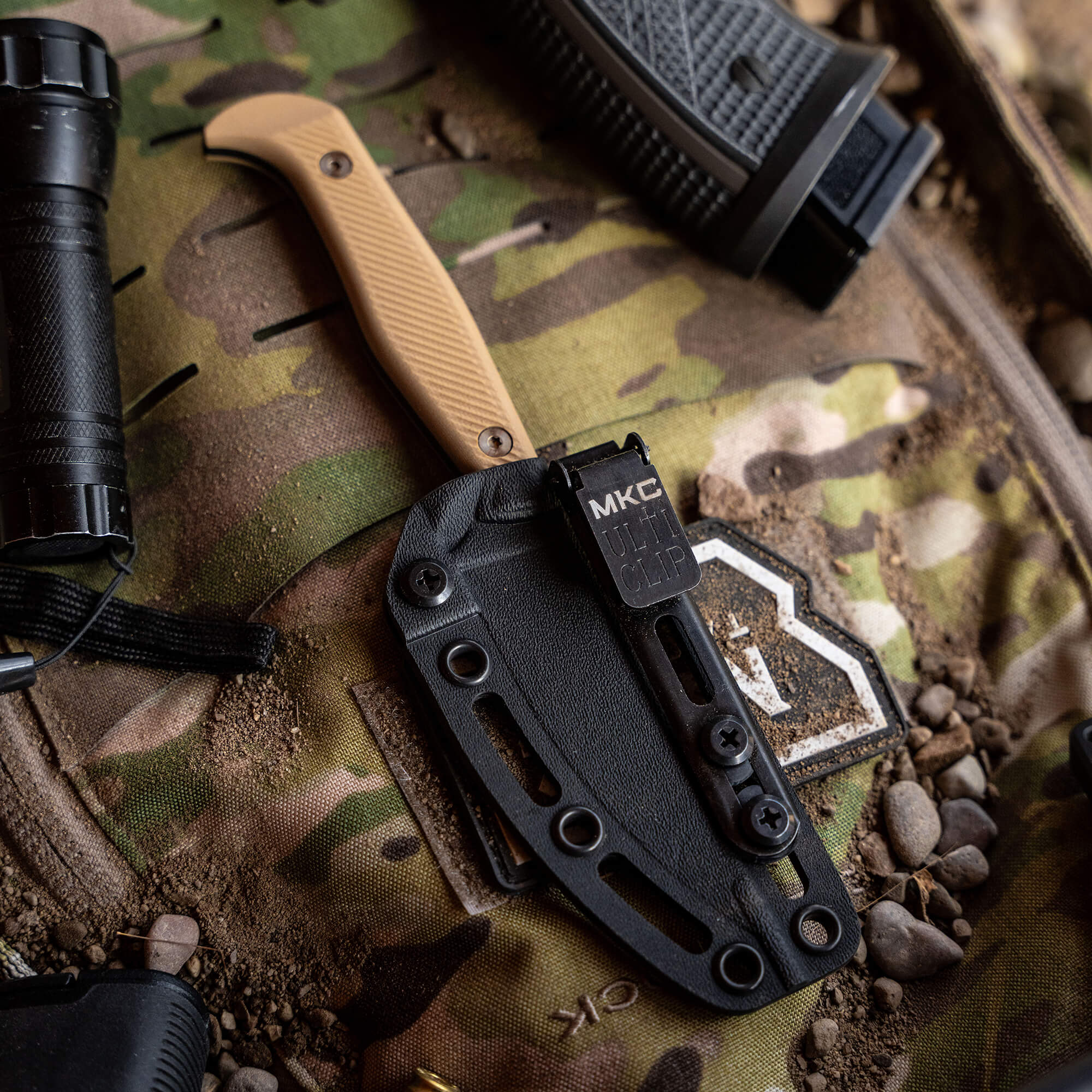 Alt text: "Close-up view of a 'Battle Goat - BLK/COY' tactical knife in a black sheath, displayed on a camouflage background with other tactical gear including another knife and a flashlight."