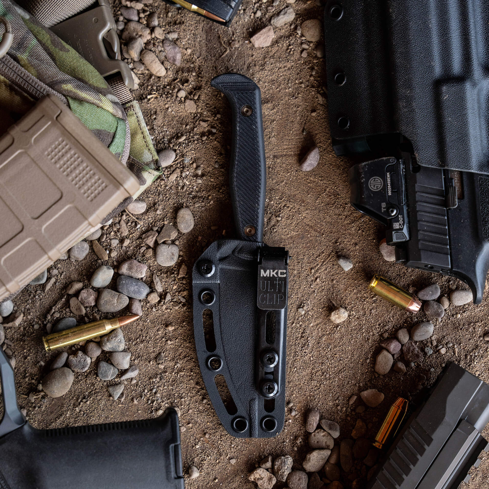 Black tactical knife with sheath, surrounded by military gear including ammunition and parts of firearms, displayed on a gravel surface.
