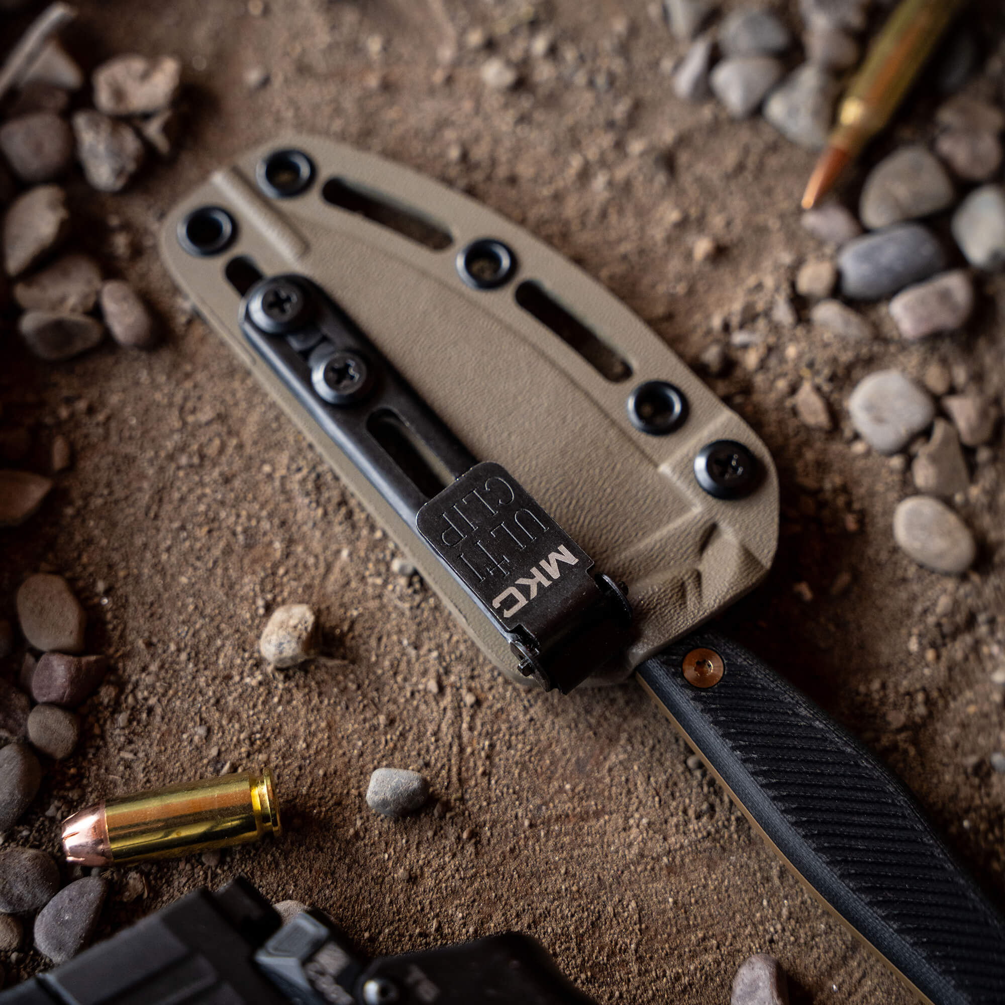 Alt Text: "Close-up of a Coyote Black Battle Goat knife in a tan sheath, resting on a gritty surface surrounded by small stones and a bullet casing."