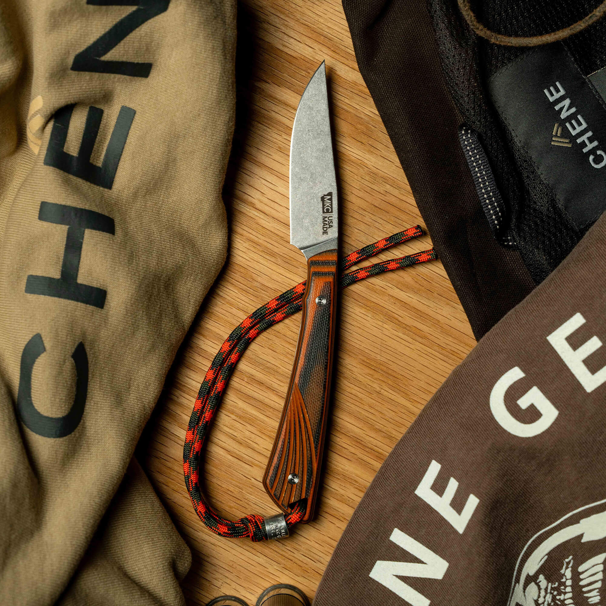 Montana Knife Company's CHÊNE Freezout knife displayed on a wooden surface with a paracord lanyard and branded apparel.