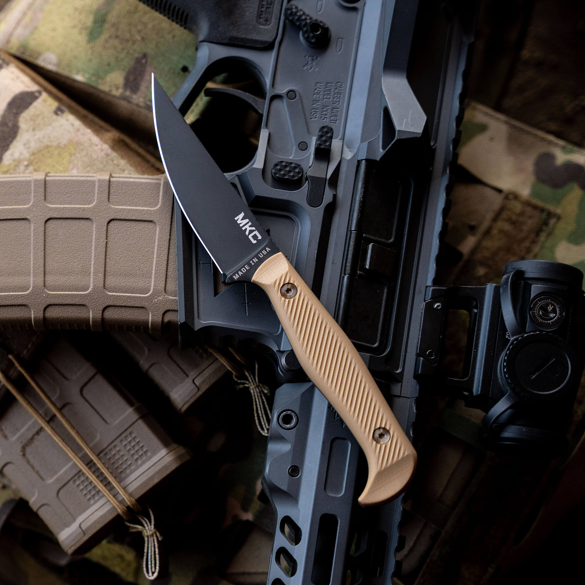 Alt text: "BATTLE GOAT tactical knife with black blade and beige handle, displayed on top of assorted tactical gear including magazines, a rifle, and a camera, all set against a camouflage backdrop."