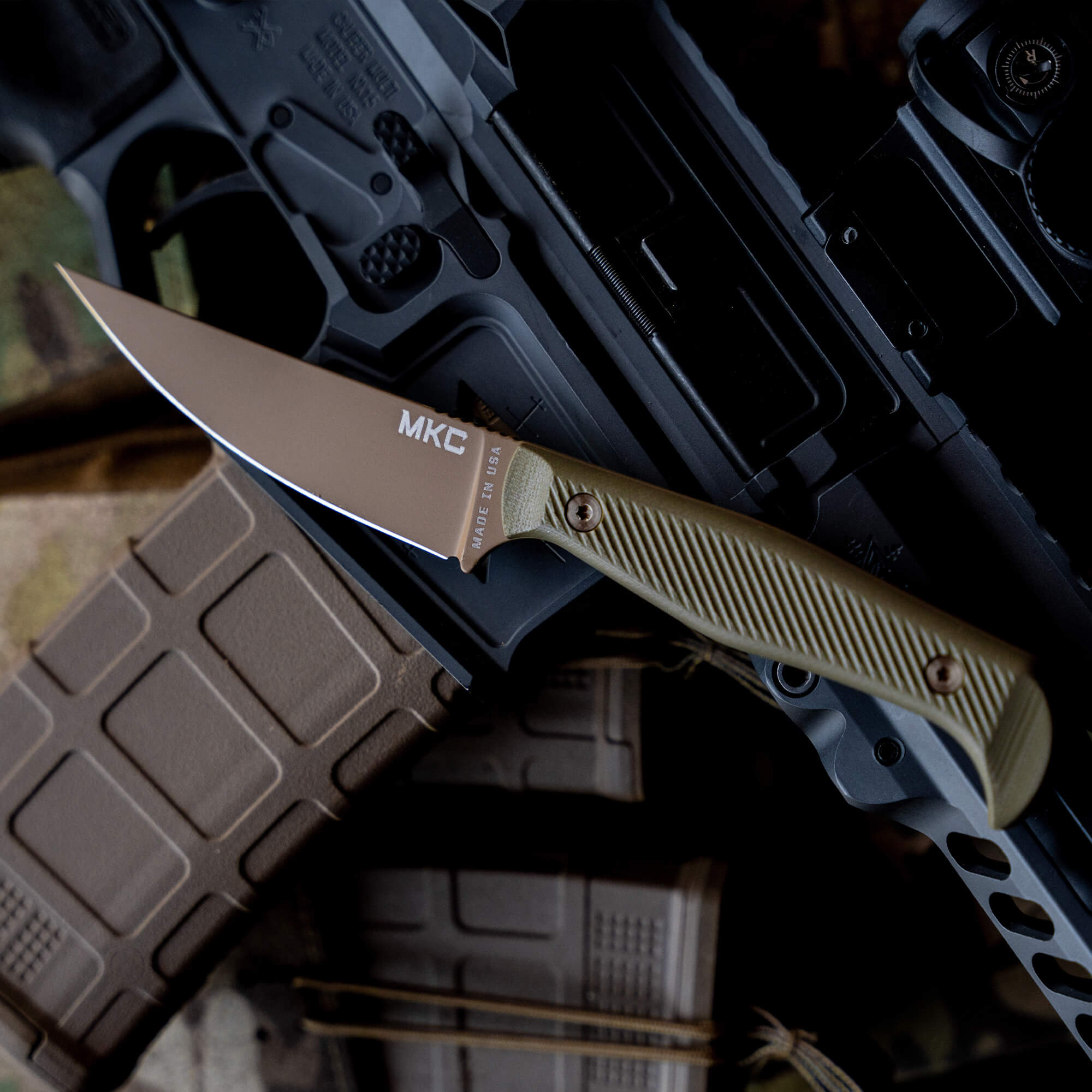 Alt text: "Close-up of the Battle Goat fixed blade knife with a coyote tan and olive drab handle, shown resting on a tactical rifle with magazines. The blade is prominently branded with 'MKC' and 'Made in USA'."
