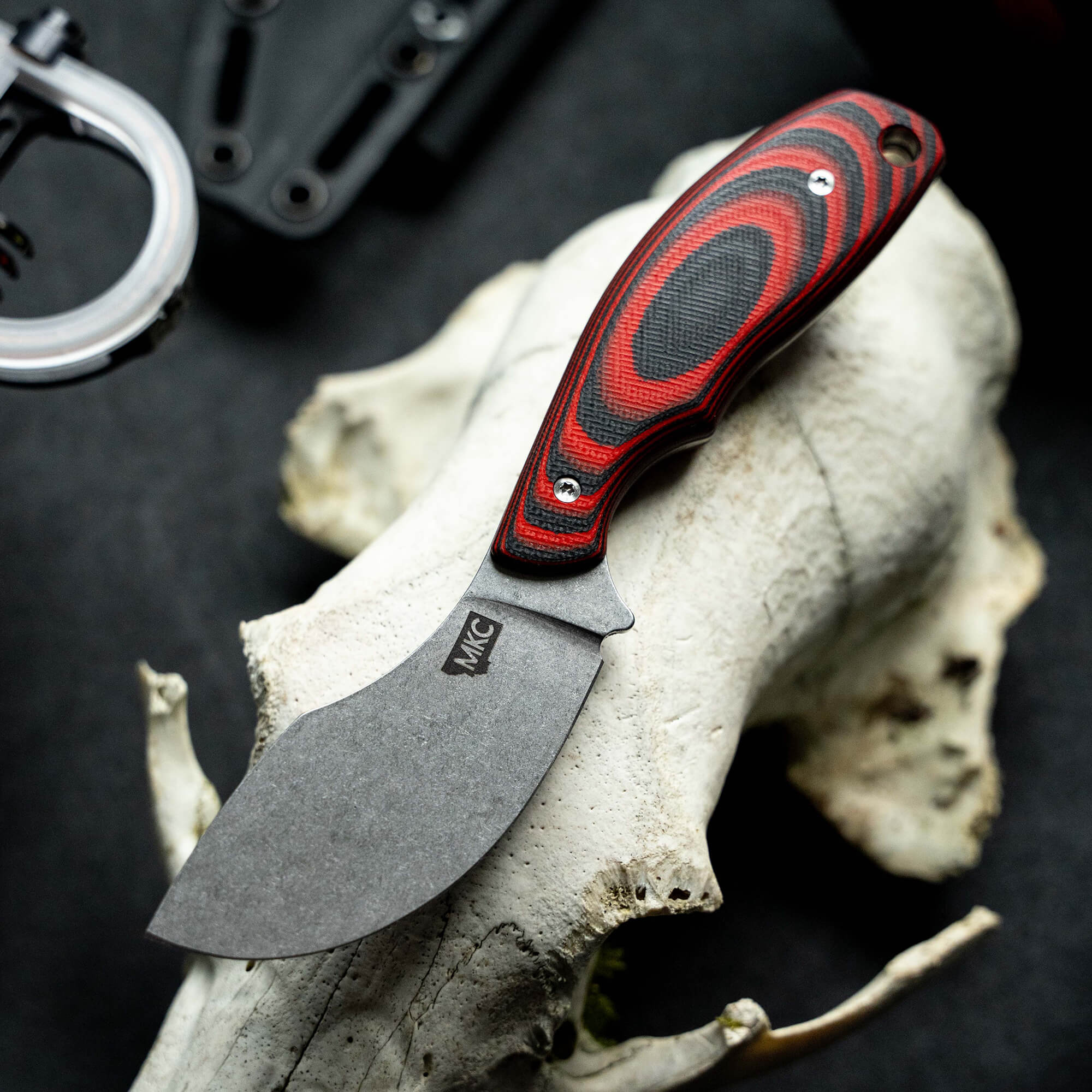 A close-up image of a fixed blade skinner knife with a red and black handle featuring the logo "MKC", displayed atop a weathered animal skull, emphasizing the ruggedness and quality of the knife.