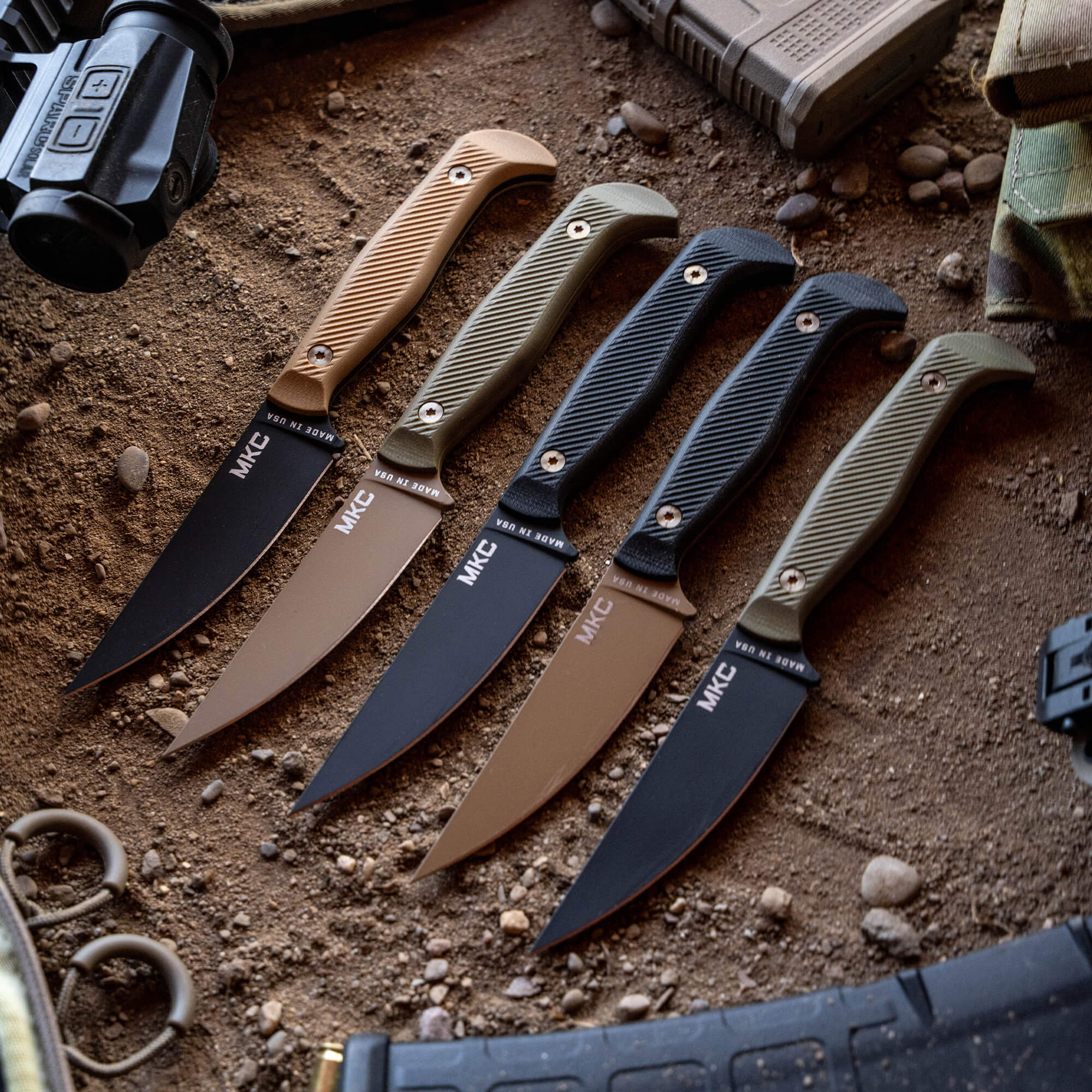Alt text: A collection of tactical knives displayed on a sandy surface, featuring various handles in coyote and black colors. The knives have a sleek design with their model name "MKE" marked on the blade.