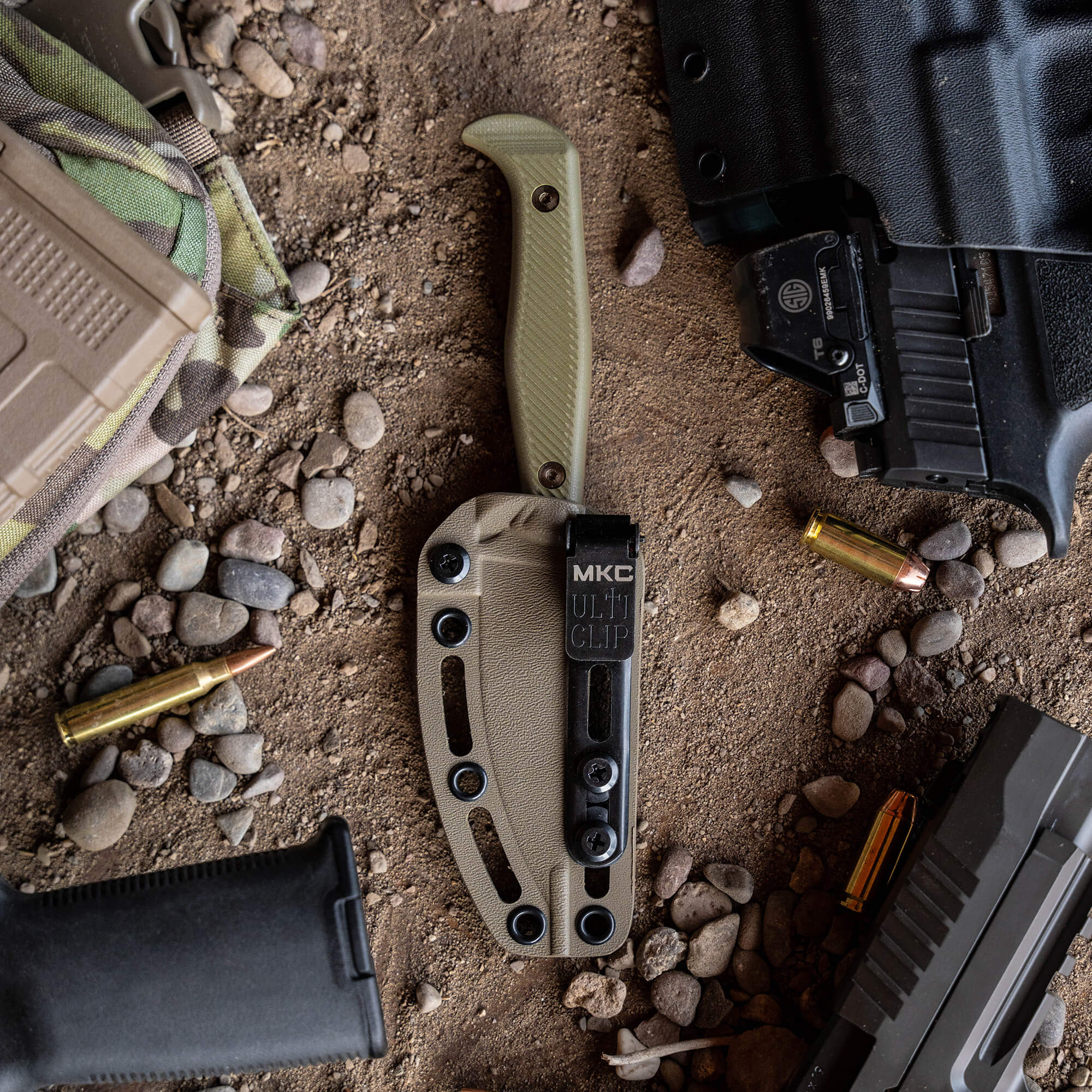 Alt text: "Close-up view of a coyote tan 'Battle Goat' tactical knife in sheath with MKC Ulti Clip, surrounded by tactical gear including a firearm, ammunition, and rugged outdoor equipment, set against a gritty, pebb