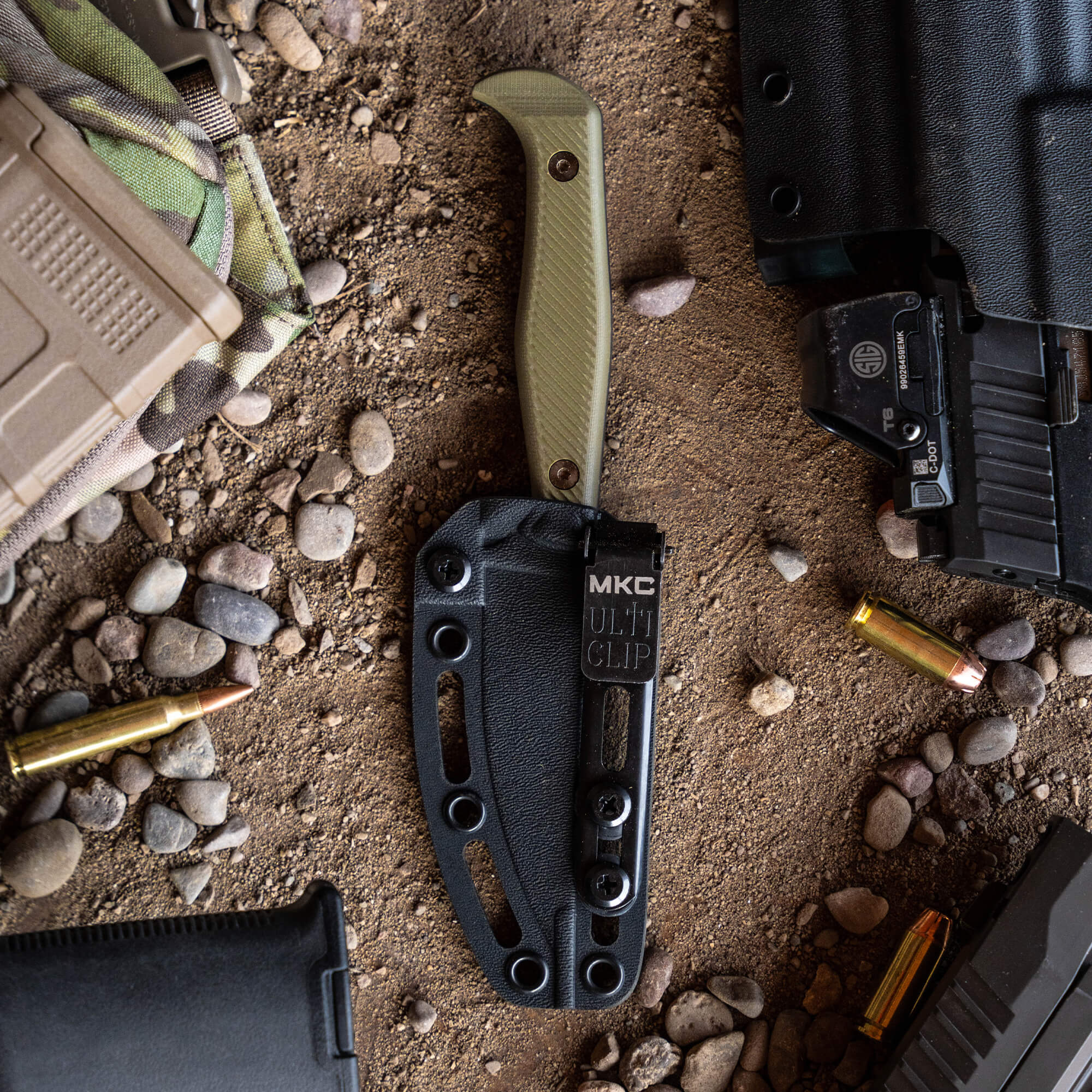 Alt text: "Close-up view of a Battle Goat tactical knife in black and olive drab with a textured handle and black sheath, displayed alongside tactical gear and ammunition on a gritty surface."