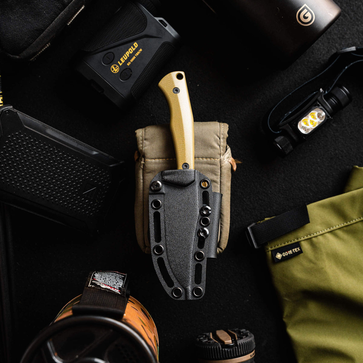 Olive TRIUMPH PRO knife with a black sheath, surrounded by outdoor gear including a flashlight and water bottle.