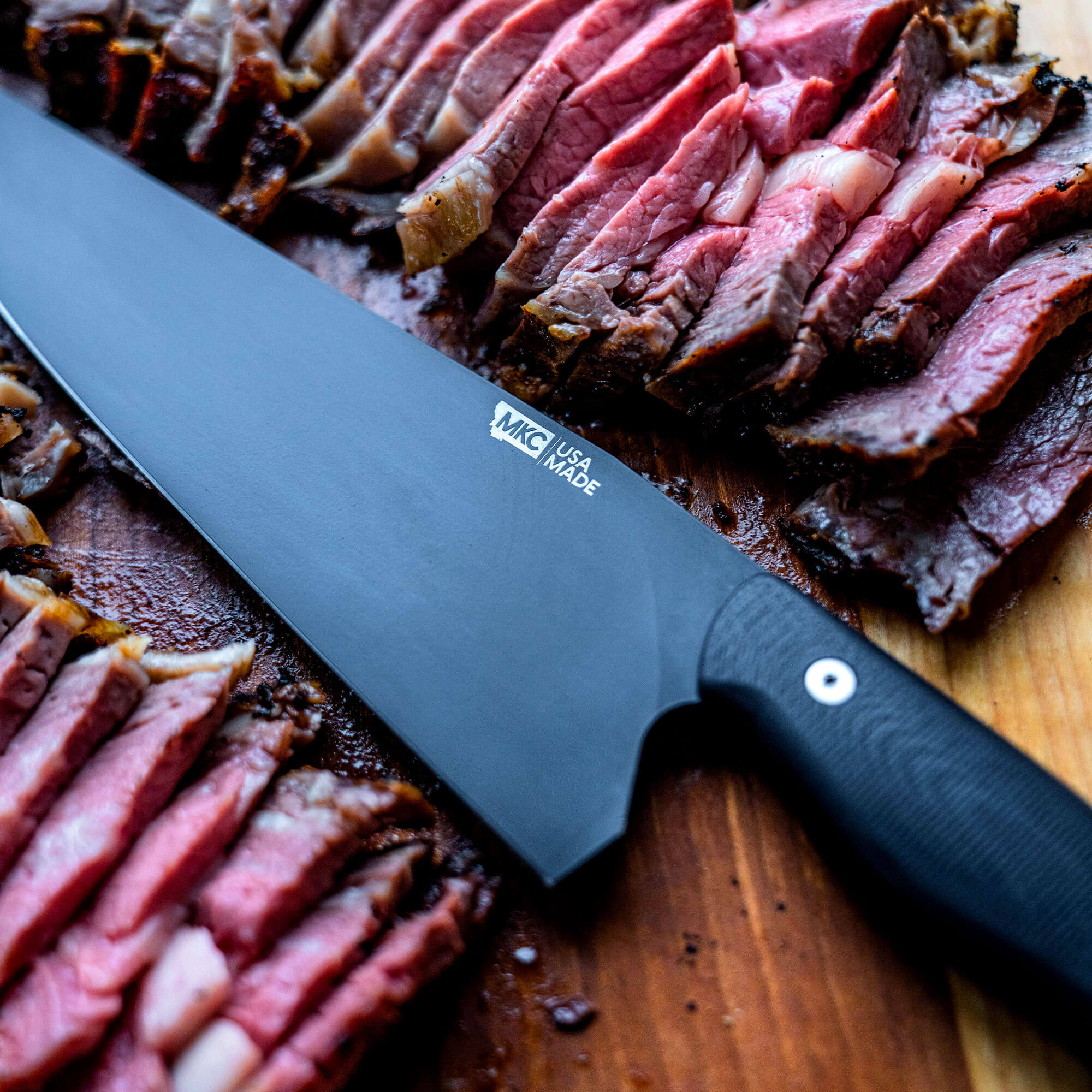 Alt text: A Bighorn Chef black kitchen knife placed on a wooden cutting board next to sliced medium-rare steak, highlighting the knife's sharp blade and ergonomic handle with logo "MKC USA Made".