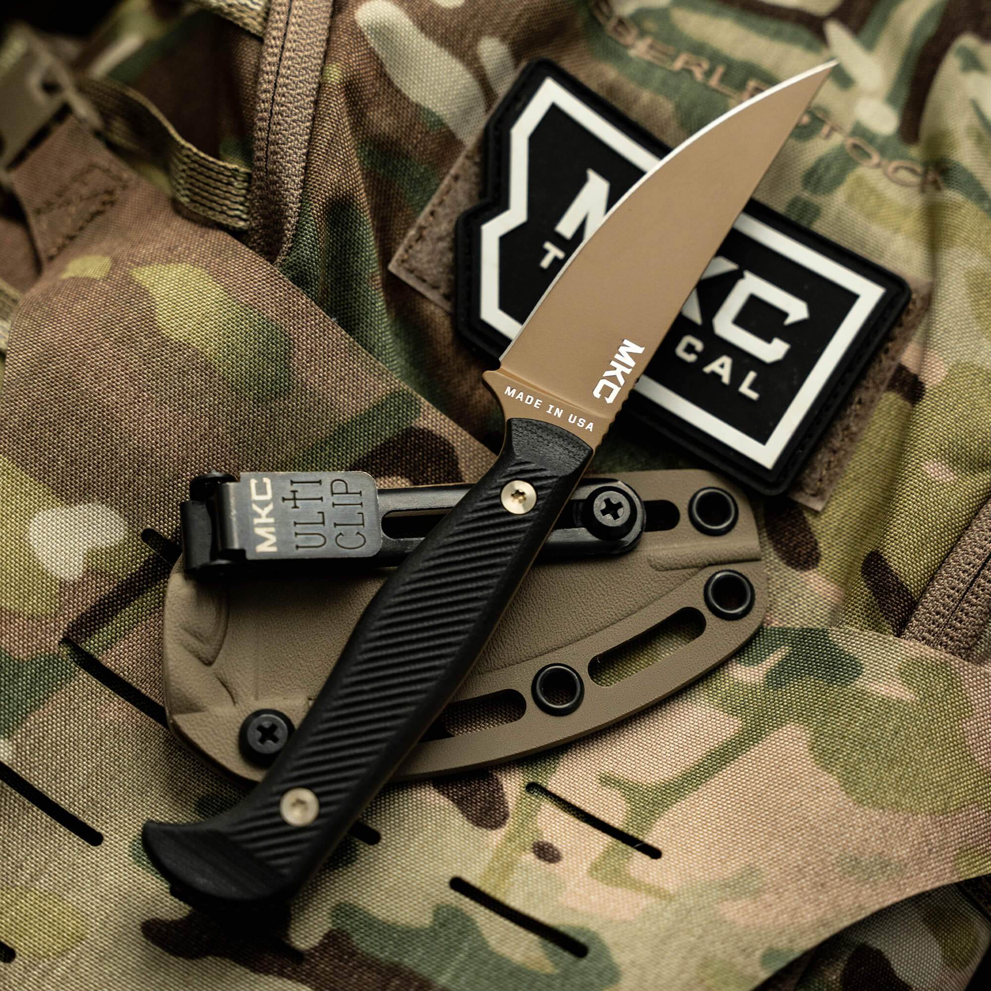 Fixed-blade knife with black handle and coyote tan blade displayed on camouflaged fabric background with tactical gear elements and brand patches, emphasizing rugged outdoor utility.