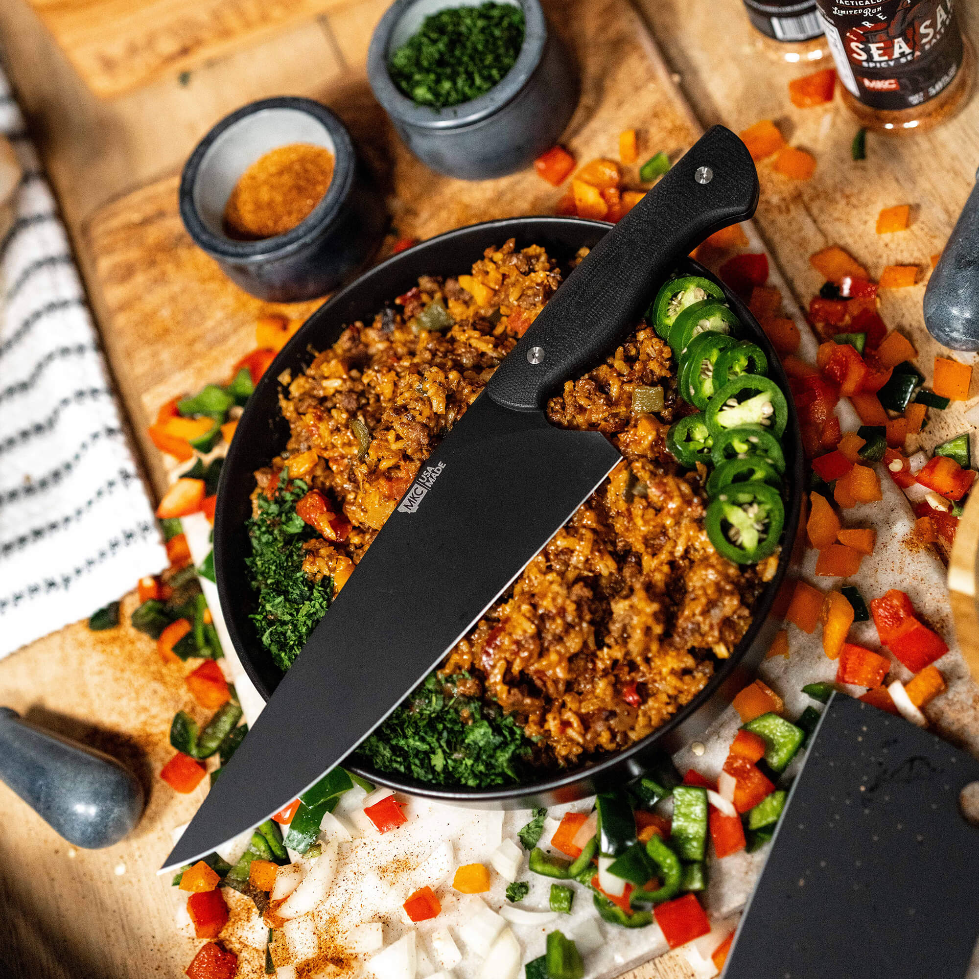 Alt text: "BIGHORN CHEF Black Series chef's knife on a wooden cutting board, slicing through a spicy minced meat dish in a skillet surrounded by chopped vegetables including bell peppers and jalapeños, with condiment bowls and seasoning containers visible