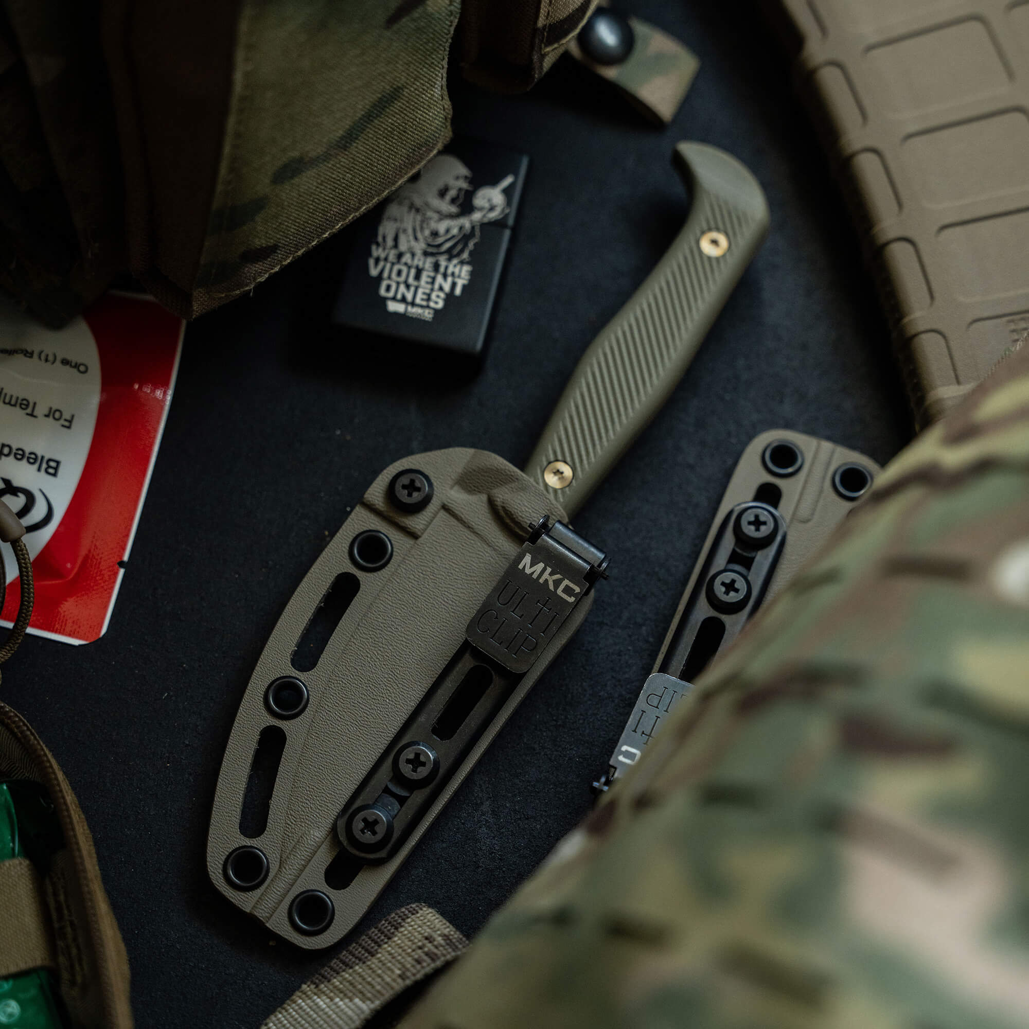 Alt text: "Tactical folding knife with olive drab handle and matching sheath amid a thematic military set-up including camouflage patterns and tactical gear accessories."