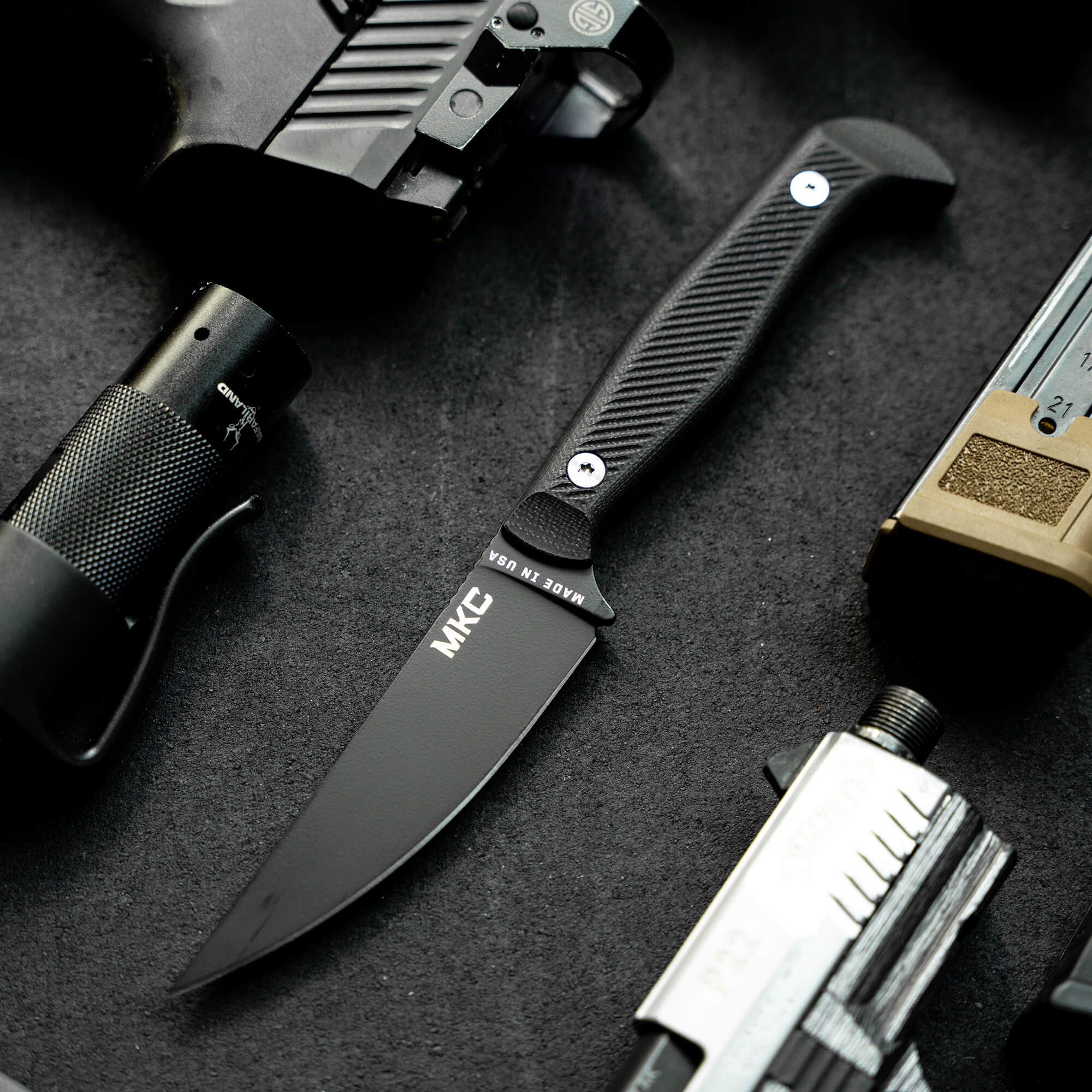 Alt text: Black tactical knife with textured handle and matte blade, branded "MKC", displayed among other tactical gear including a flashlight and measuring tool, set on a dark textured surface.