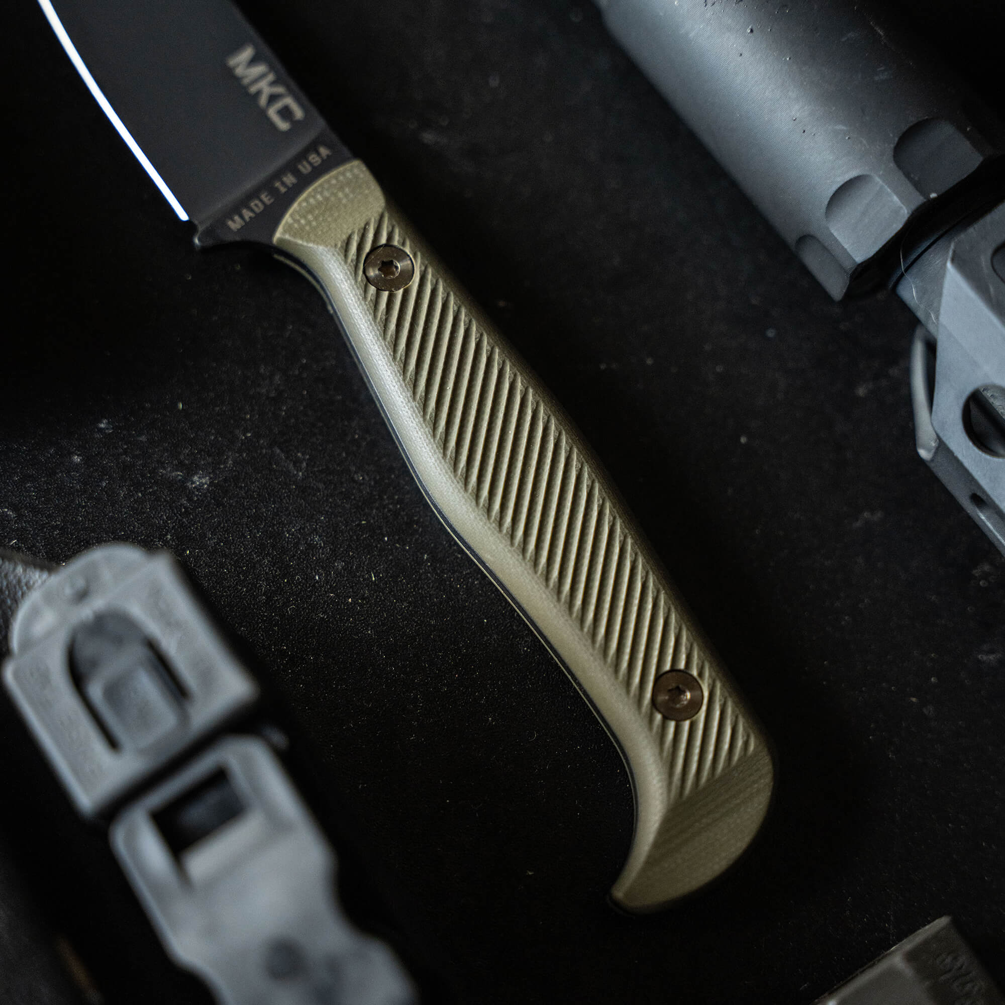 Close-up of a BATTLE GOAT tactical knife with a black blade and olive drab handle, displayed alongside tactical gear.