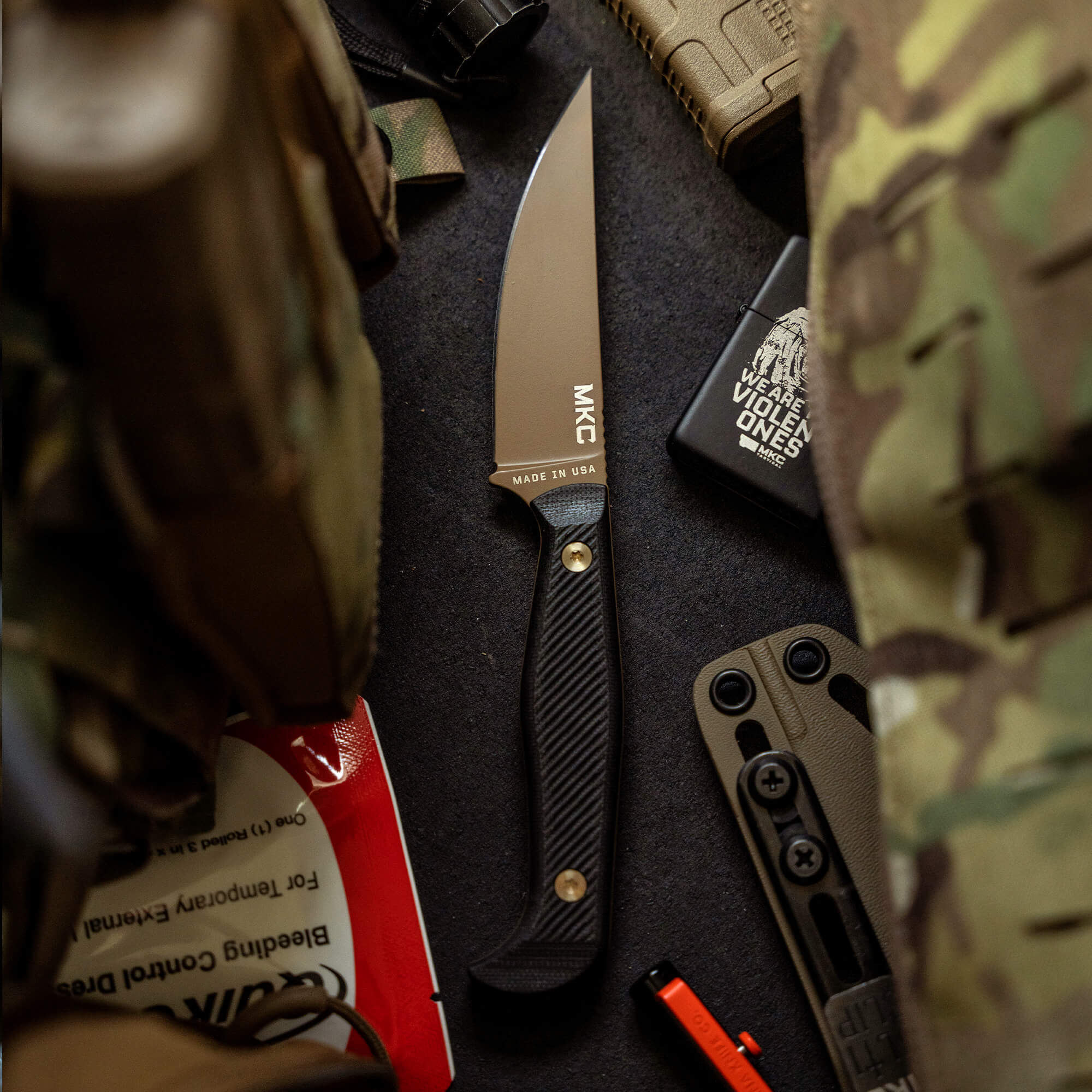 Alt text: "Close-up of the MKC Battle Goat knife in Coyote Black, featuring a sleek blade and a textured handle, displayed alongside tactical gear including a camo pattern backpack, boots and various survival tools."