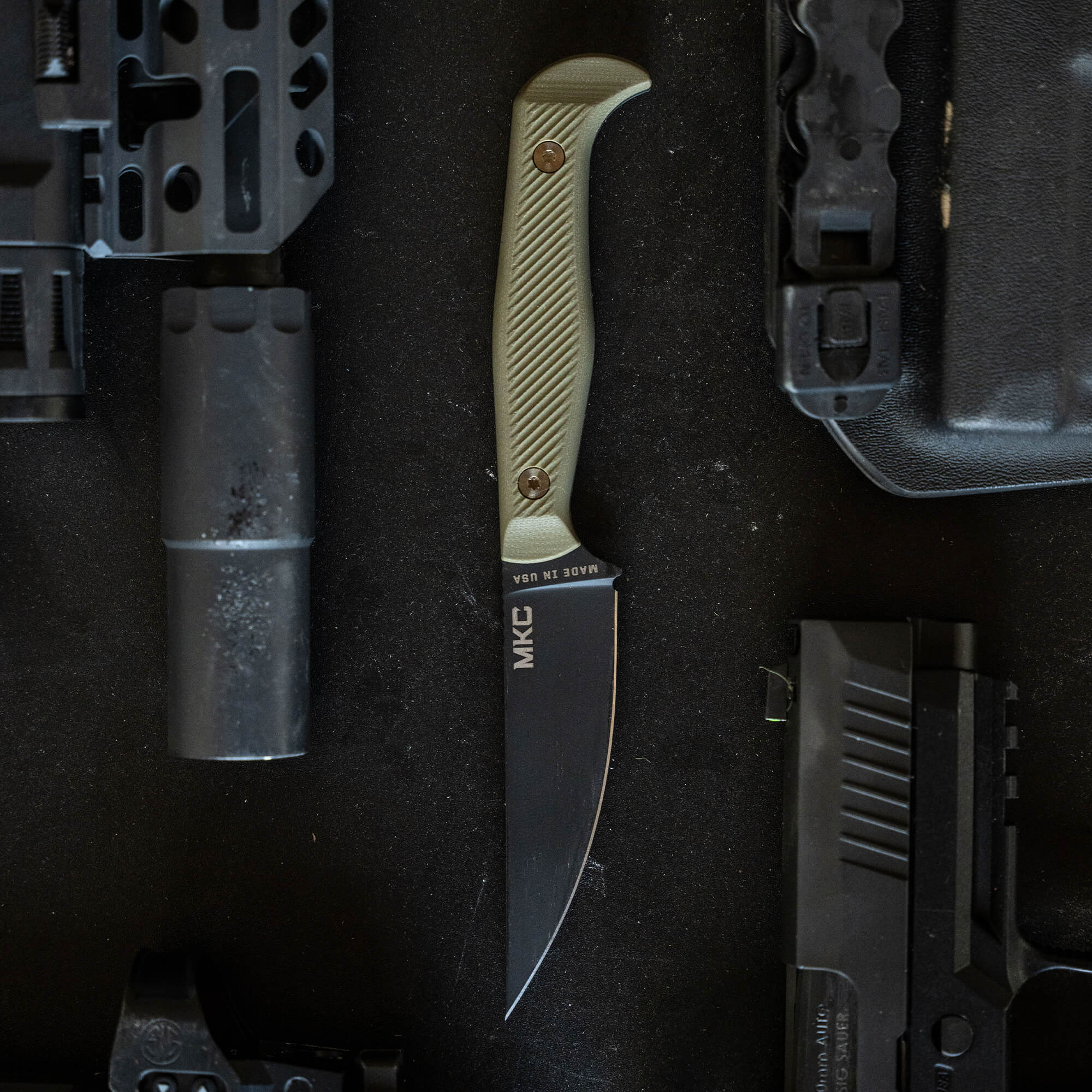 A tactical knife named "BATTLE GOAT - BLK/OD" with a beige textured handle and black blade, marked "MKC," displayed alongside gun parts and equipment on a dark, textured surface.