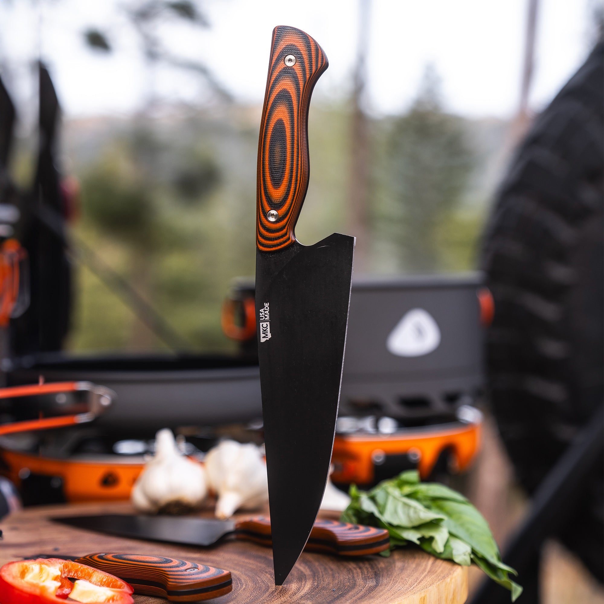 Alt text: "Bighorn Chef heavy-duty orange and black camping knife with a uniquely patterned handle, prominently displayed in an outdoor cooking environment, with blurred cooking utensils and nature in the background."