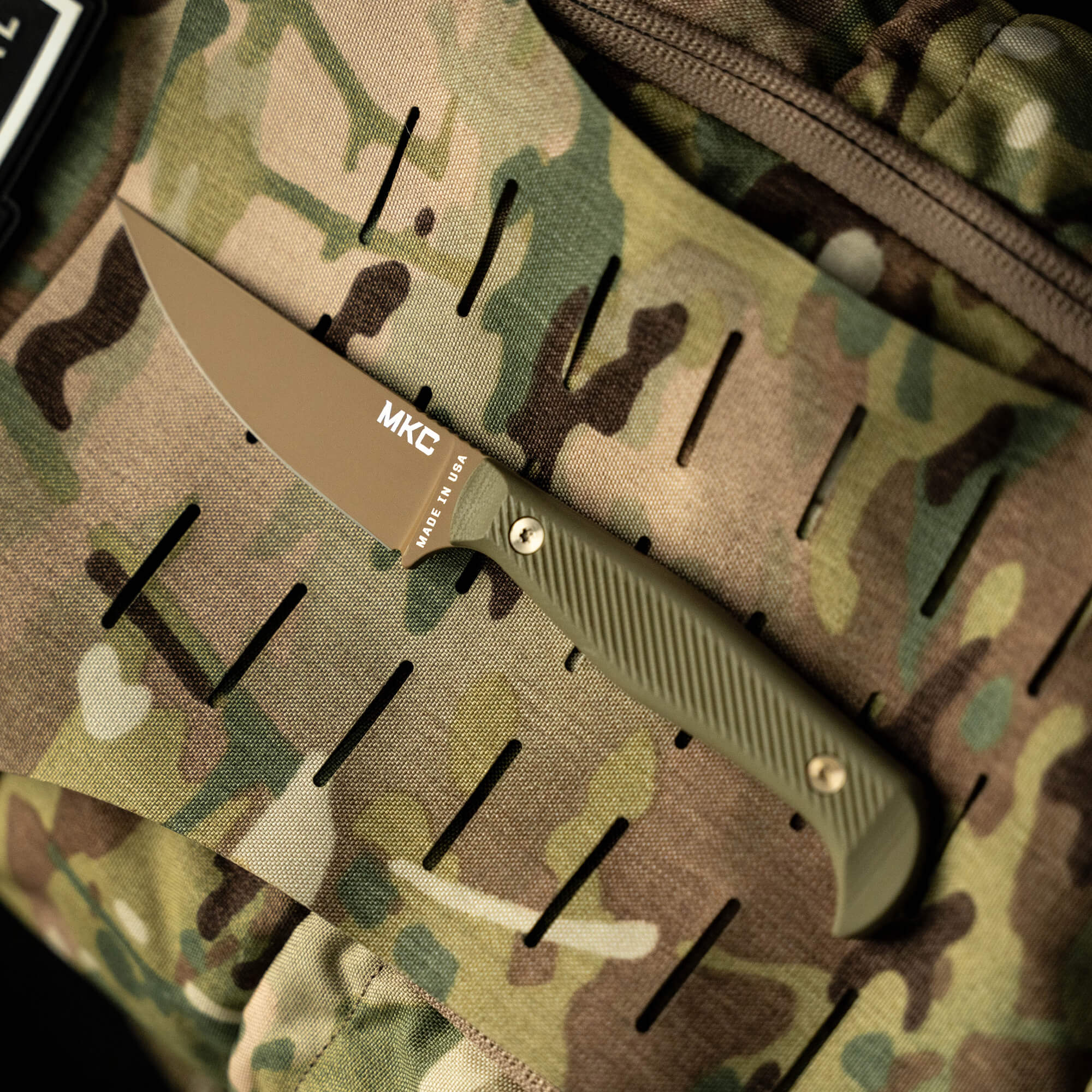 A tactical knife with a tan blade and olive green handle, branded 'MKE', displayed against a camouflage background.